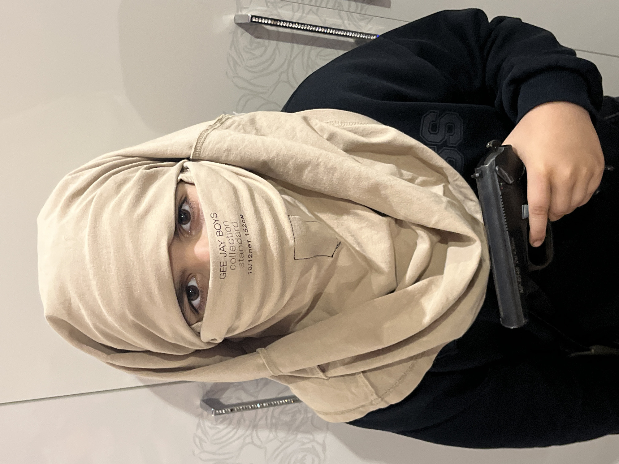 A young person, possibly a child, is shown wearing a beige headscarf that covers their hair, neck, and the lower half of their face. The headscarf has a label with text that appears to say "GEE JAY BOYS collection standard 10/12лет 152см". The person is also wearing a dark blue hooded sweatshirt. In their right hand, the person is holding a black handgun. The background appears to be an interior setting, possibly a room in an apartment in Kostroma, Russia. The walls are light-colored and have a subtle patterned texture. A portion of a wardrobe or cabinet is visible on the left side of the image, featuring a vertical metallic handle with embedded rhinestones. Another similar decorative strip is visible on the right side of the image. The lighting suggests indoor illumination.