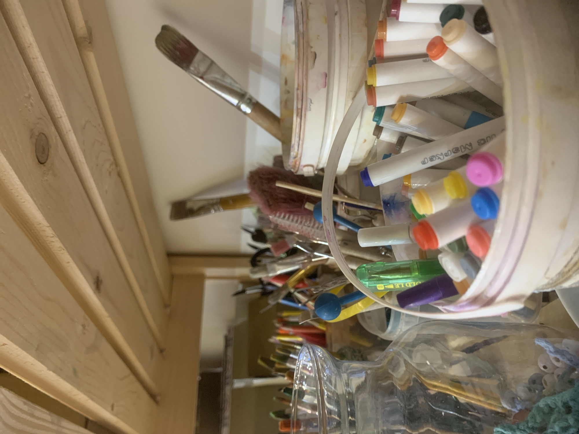 The image depicts a cluttered art supply storage area. A set of unfinished wooden shelves is visible on the left side of the frame. The shelves hold a variety of art supplies, including brushes, markers, and other tools.
A white plastic bucket filled with markers dominates the center of the image. The markers have white barrels and colorful caps.
In the lower right corner of the image, a clear glass jar is visible. It appears to contain small objects, possibly beads or other craft supplies.
Given the cluttered nature of the scene and the presence of art supplies, it is likely that this image was taken in an artist's studio or workspace. There is no visual information in the image that points specifically to the city of Izhevsk, Russia.