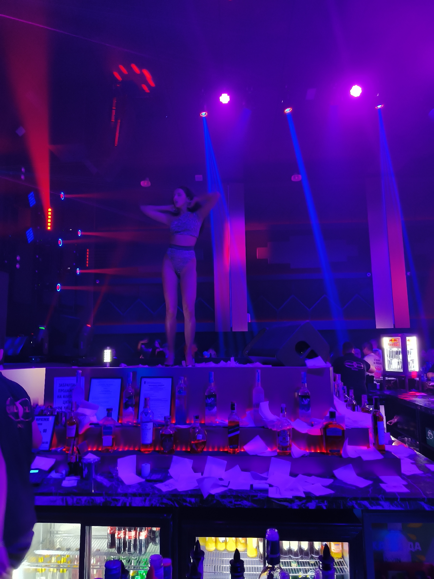 In Kiten, Bulgaria, a woman in a bikini stands on a stage under red and blue lights. She is posing with her hands behind her head. The stage is decorated with bottles and papers, and there are speakers and lights in the background. The scene appears to be in a nightclub or bar.