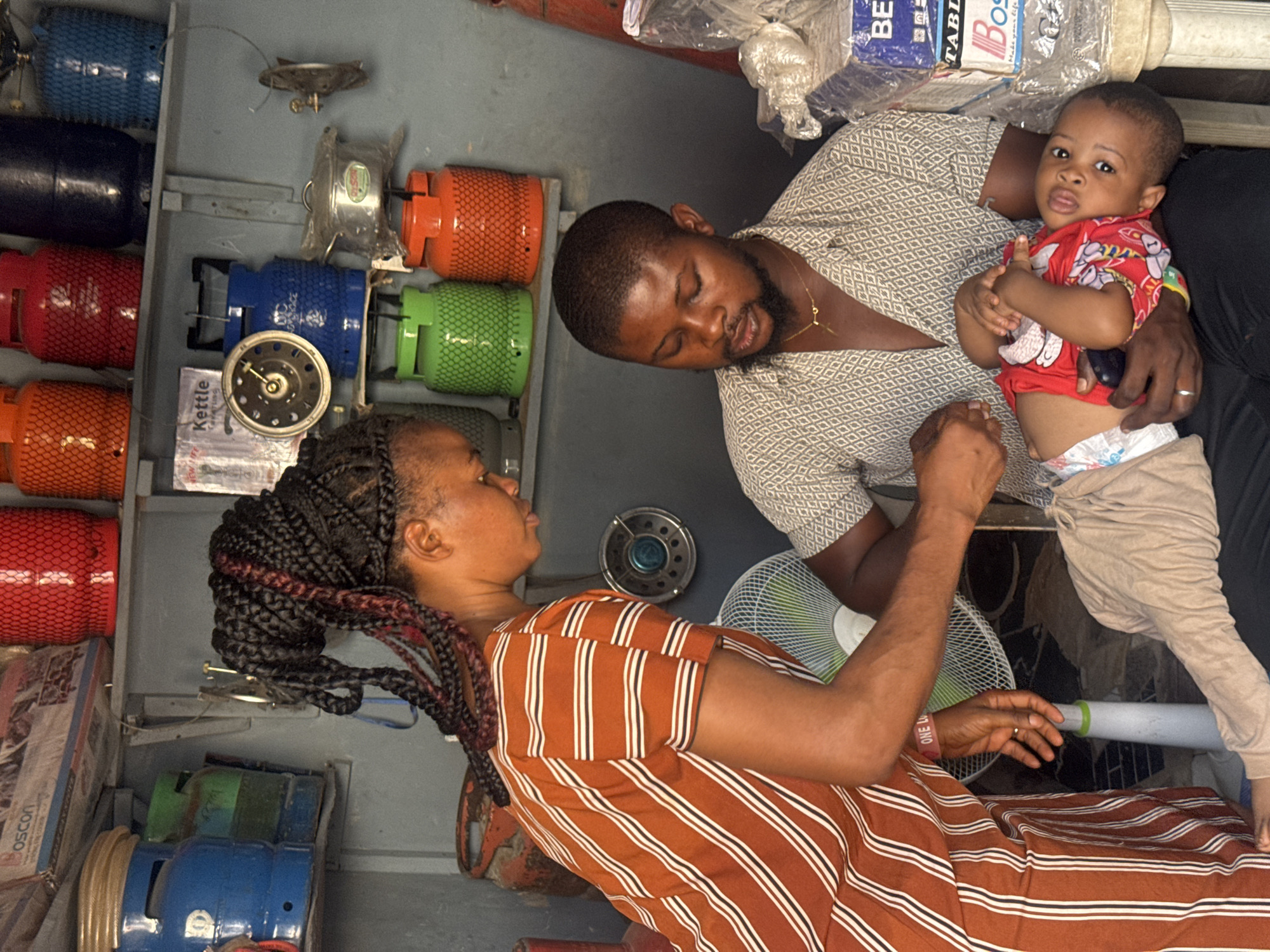 This image captures a family scene within what appears to be a shop or storage facility for gas cylinders and related appliances in Ihiagwa, Nigeria. The left side of the frame features shelves densely packed with numerous LPG cylinders of various colors including red, orange, blue, green, and black. Several gas cookers, some still packaged, are also visible, along with a box labeled "Kettle" and another box with "OSCON" partially visible.
In the center, a man, likely in his late 20s or early 30s, with a beard and wearing a patterned short-sleeved shirt and a gold chain, is holding an infant. The baby, dressed in a red patterned t-shirt and light-colored trousers over a diaper, looks directly towards the camera with a curious expression. The man wears a silver ring on his left ring finger and looks down gently at the baby.
Opposite them stands a woman, also appearing to be in her late 20s or early 30s, with braided hair featuring some reddish strands. She is wearing a brown and white striped dress or top and has her right hand extended towards the man and baby, her gaze fixed on them in what seems to be an attentive or engaging manner. She wears a bracelet on her right wrist that clearly reads "ONE LOVE" and a silver ring on her left ring finger. A white desk fan is visible near her right arm.
Above the man and baby, various packaged items are stacked, including bags with partially visible text such as "BETTABLE" and "BOO...". The overall scene portrays a familial interaction within an indoor setting, likely a commercial space given the prominent display of merchandise. The lighting suggests it is an indoor environment, possibly during the day.