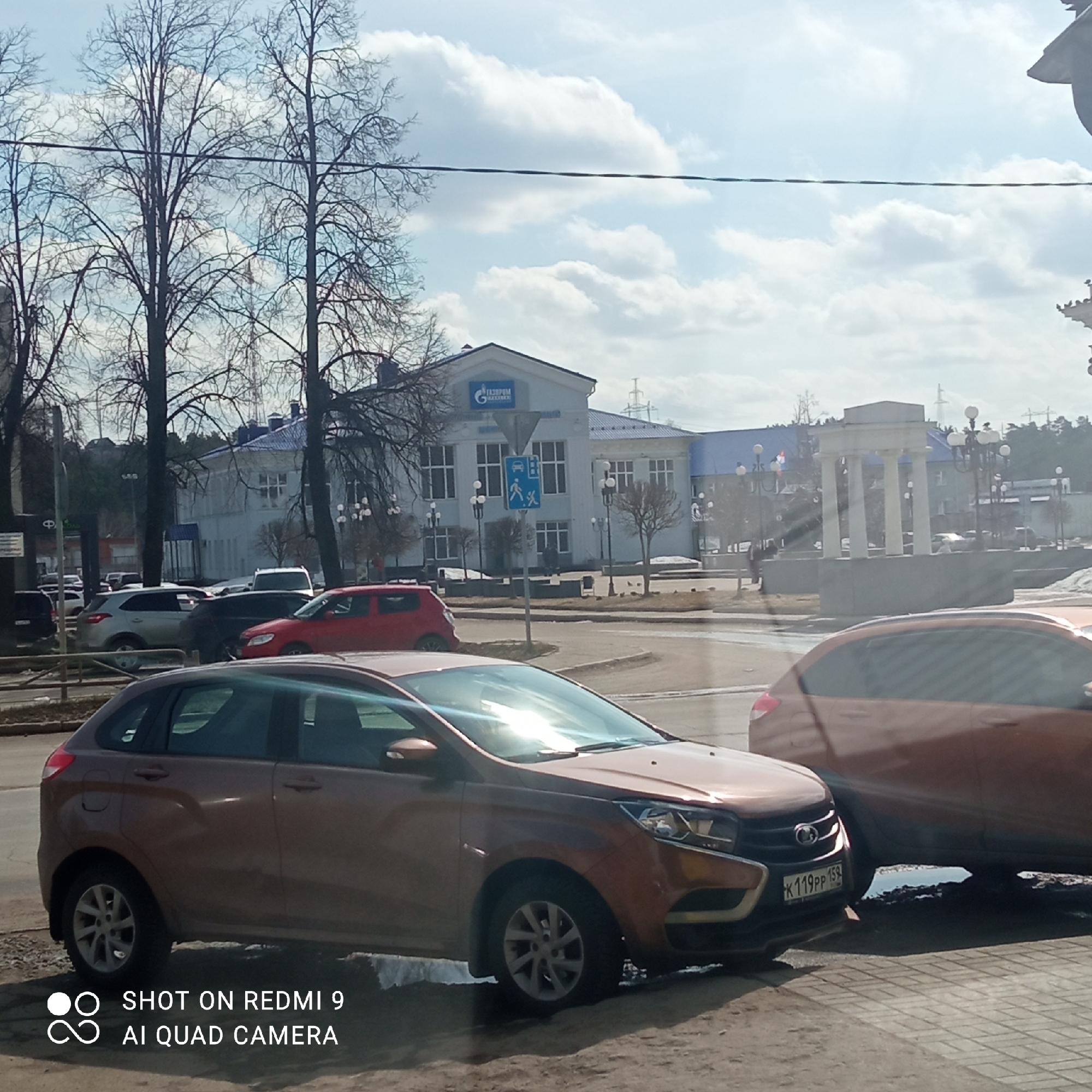 The image depicts an urban scene in Chaikovsky, Russia, on a day with a partly cloudy sky. In the foreground, there are two bronze-colored Lada XRAY cars parked near a curb. The nearest vehicle has a license plate that reads "K119PP 159." Behind the cars, there is a traffic circle with a variety of other vehicles visible. A building with the Gazprom logo on its facade can be seen in the background, along with trees and a white classical pavilion-like structure with columns. Pedestrian crossing and public transport signs are visible near the Gazprom building. Bare tree branches and overhead power lines are also present. The overall setting appears to be a relatively quiet urban area with mixed architectural styles.