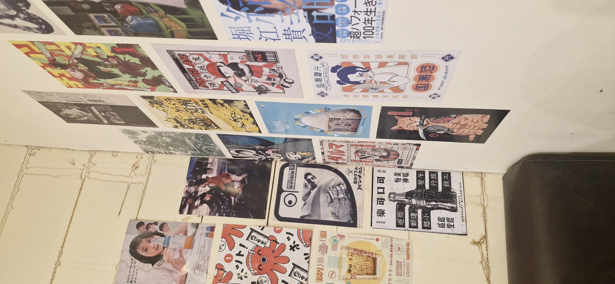 The image shows a wall covered with various posters. On the left, there is a poster of a woman holding a bottle of Fanta. To its right, there are posters with Japanese text and illustrations, including one with an octopus design and another with a vintage-style advertisement. In the center, there are more posters with a mix of Japanese text, illustrations of people, and abstract designs. One poster features a black and white image of a man. Towards the right, there are posters with vibrant illustrations, including a character on a scooter and a woman with a fan. A poster with a cat holding a sword is visible in the lower right section. The wall itself appears to be painted in a light color and has some visible stains or markings. A portion of a dark brown leather sofa is visible in the lower left corner of the image. The overall impression is that of a display of art or advertisements on a wall, possibly in a cafe or shop. There is no indication of the specific location in Rantau, Indonesia.