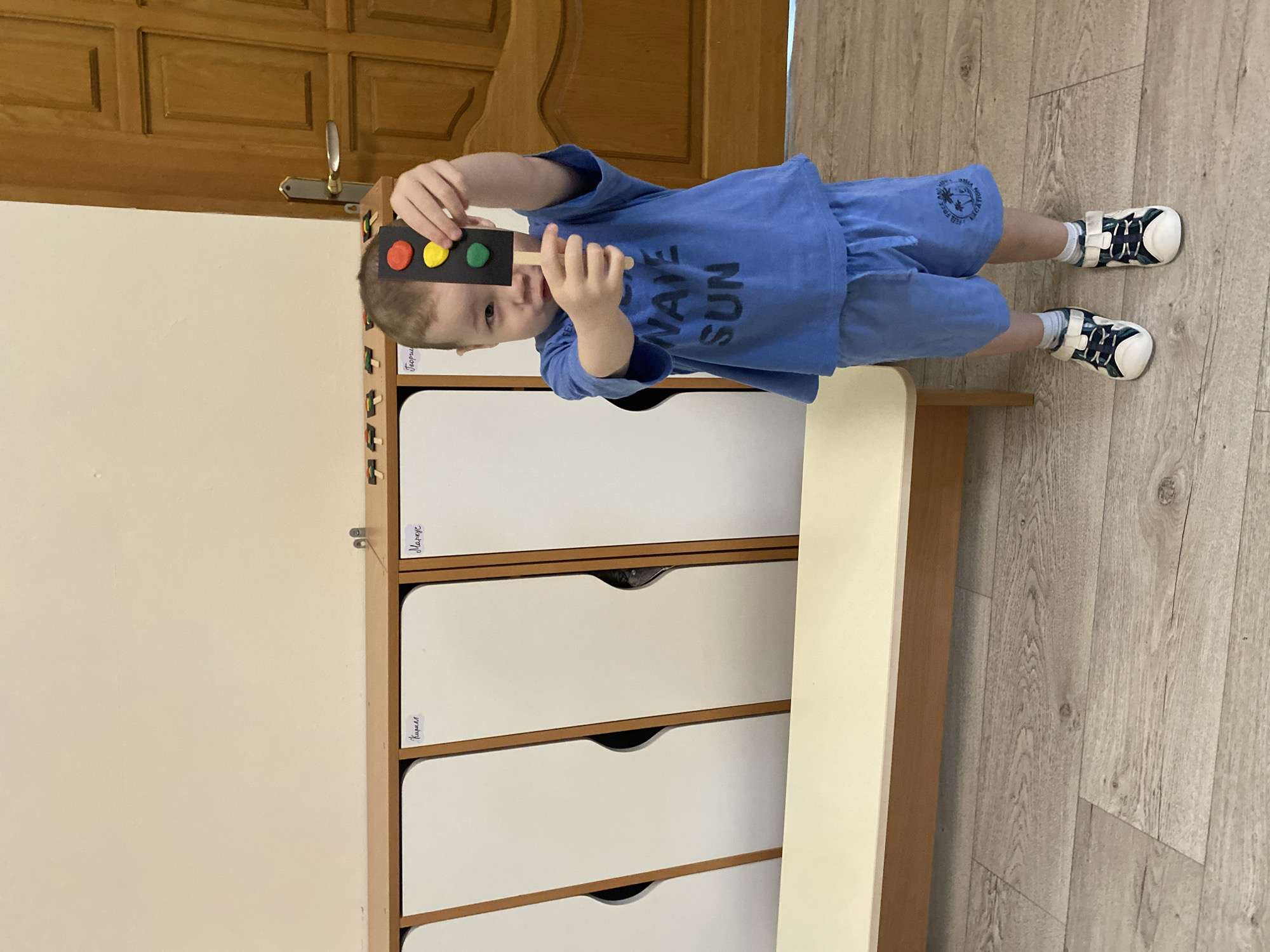 A young child, possibly around 3-4 years old, is holding up a makeshift traffic light in what appears to be a children's room. The traffic light is a black rectangle with three colored circles (red, yellow, and green) attached to it, presumably made for a game or learning activity. The child is wearing a blue t-shirt with the words "WAVE SUN" printed on it, along with blue shorts and white/blue sandals.
The child is standing on a wooden floor, near a set of white drawers. On the side of the drawers there are labels in Russian. The scene is lit with natural light.
Given the Russian labels, it is possible that this scene is in a Russian-speaking country, possibly Russia. Without further information, it's not possible to determine the location in the city of Sochi with certainty.