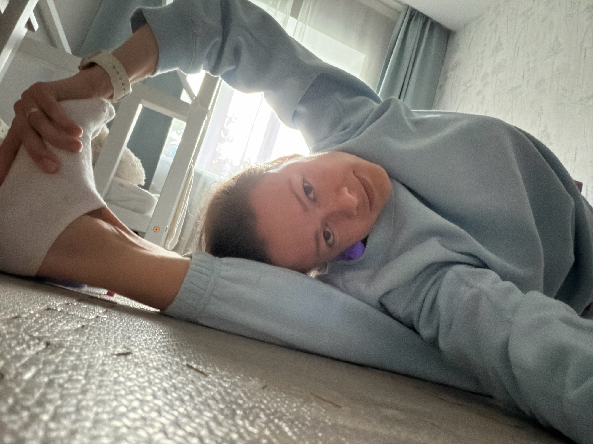 A woman is performing a split on a wooden floor. She is wearing a light blue sweatshirt and white socks. Her right leg is extended in front of her, and her left leg is bent behind her, with her left arm holding her left foot. The woman is looking at the camera with a slight smile. The background shows a window with light curtains and a wall with floral wallpaper. The scene appears to be in a residential setting, possibly a bedroom or living room. There is no indication of the location being in the city of Riga, Latvia.