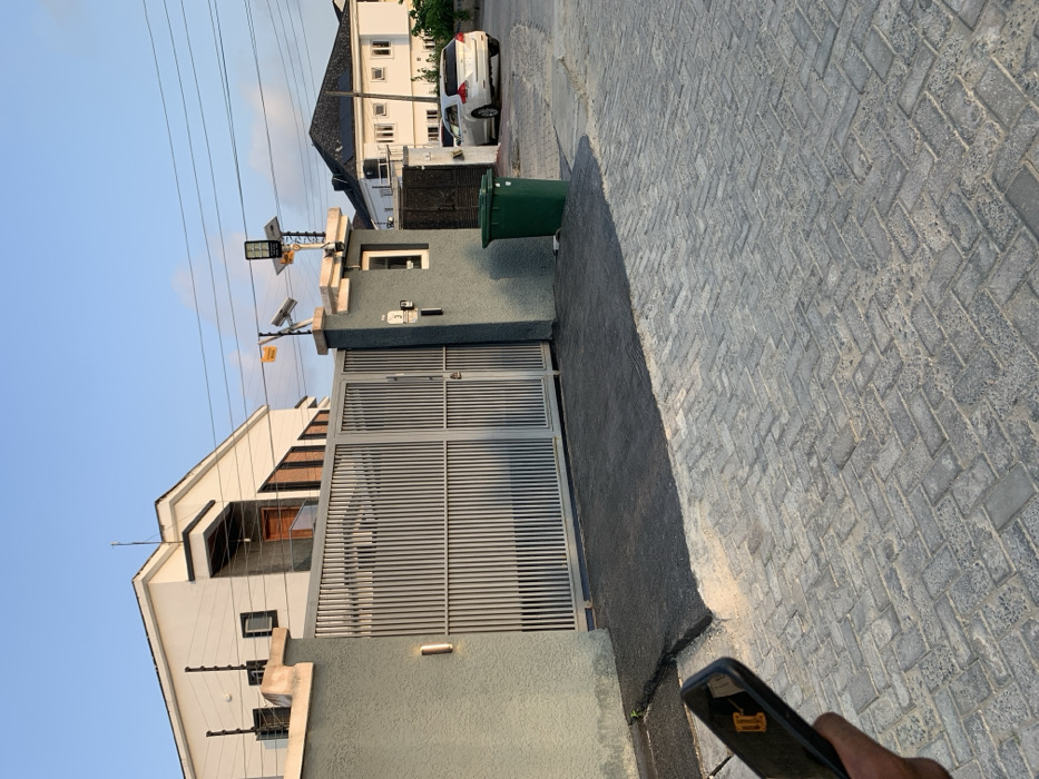 The image displays a residential street scene, rotated 90 degrees clockwise. A clear blue sky dominates the left side of the frame, with several utility wires stretched across it. The right side of the image reveals a paved ground surface, composed of grey interlocking bricks laid in a herringbone pattern, with a strip of darker asphalt-like material running parallel to a building wall.
At the center, the entrance to a modern residential property is visible. It features a large grey, slatted metal gate, including a separate pedestrian access point, set within a grey textured wall. Mounted on concrete pillars above the gate are solar-powered security lights, and what appear to be elements of an electric fence line the top of the boundary wall. A small white label near the gate displays "NO. 3," and an adjacent yellow sign partially reads "THIS PROPERTY IS PROTECTED BY...". A green trash bin is positioned next to the gate, closer to the paved area.
A portion of the main house, characterized by its white upper structure and brown-accented balconies, is visible to the upper-left of the gate. In the background, along what would be the street, another multi-story white residential building with a dark roof is seen, with a white car parked in front. In the bottom-right corner, a person's hand holds a black smartphone, on the back of which a yellow sticker shows partial text, including "Commence" and "2017." The long shadows cast across the paved ground suggest the time of day is late afternoon or early evening under clear, sunny weather. The scene is consistent with a residential area in Ikota, Nigeria.