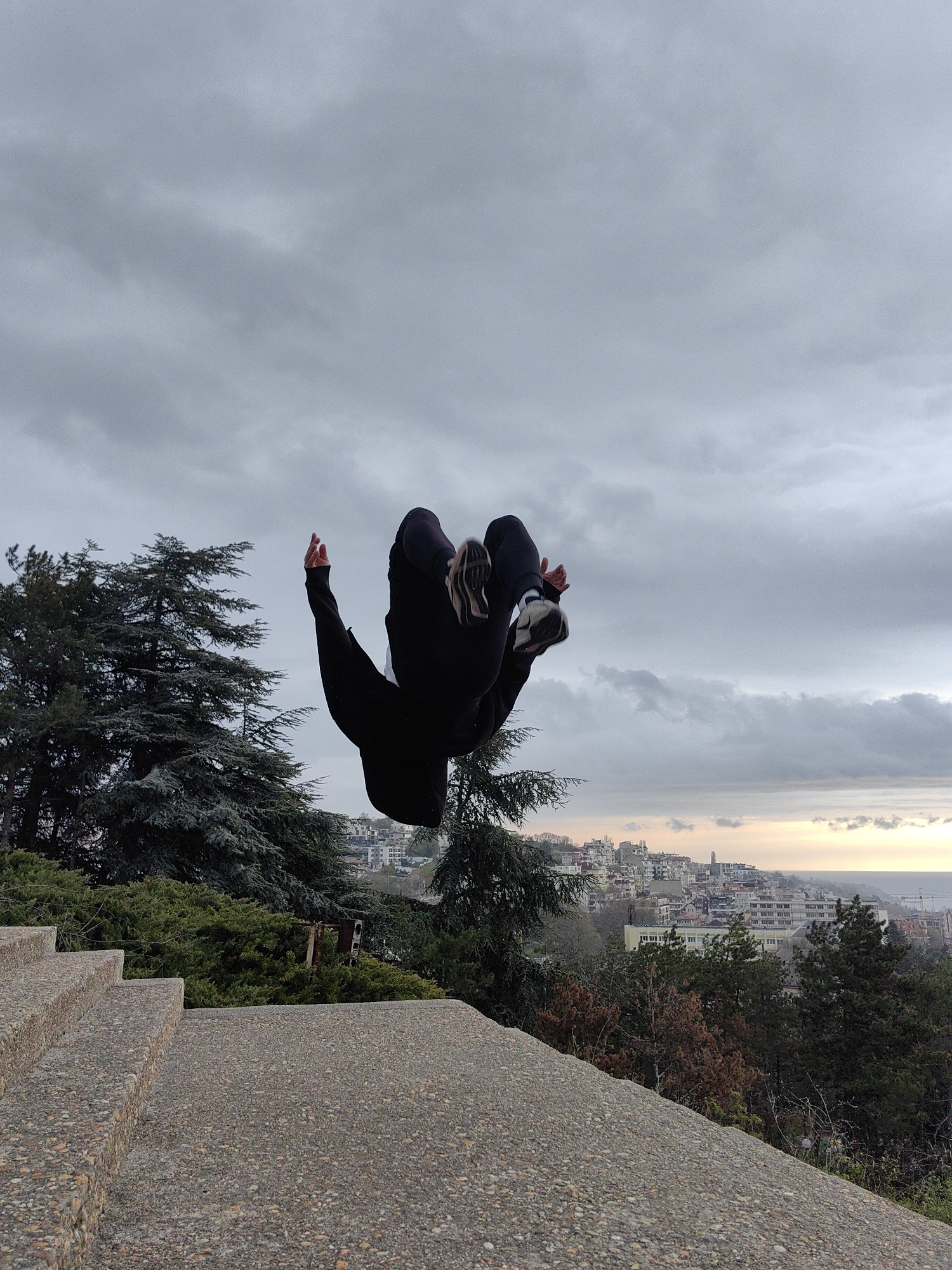 The image depicts a person in mid-air performing a backflip. The person is wearing black clothing and white sneakers. Their arms are extended upwards, and their body is inverted with their head pointing downwards. The background consists of a cloudy sky, trees, and a cityscape. The cityscape features buildings and the sea. The immediate foreground shows concrete stairs and a ledge. The location is Varna, Bulgaria, specifically the Seaside Park, overlooking the Black Sea.