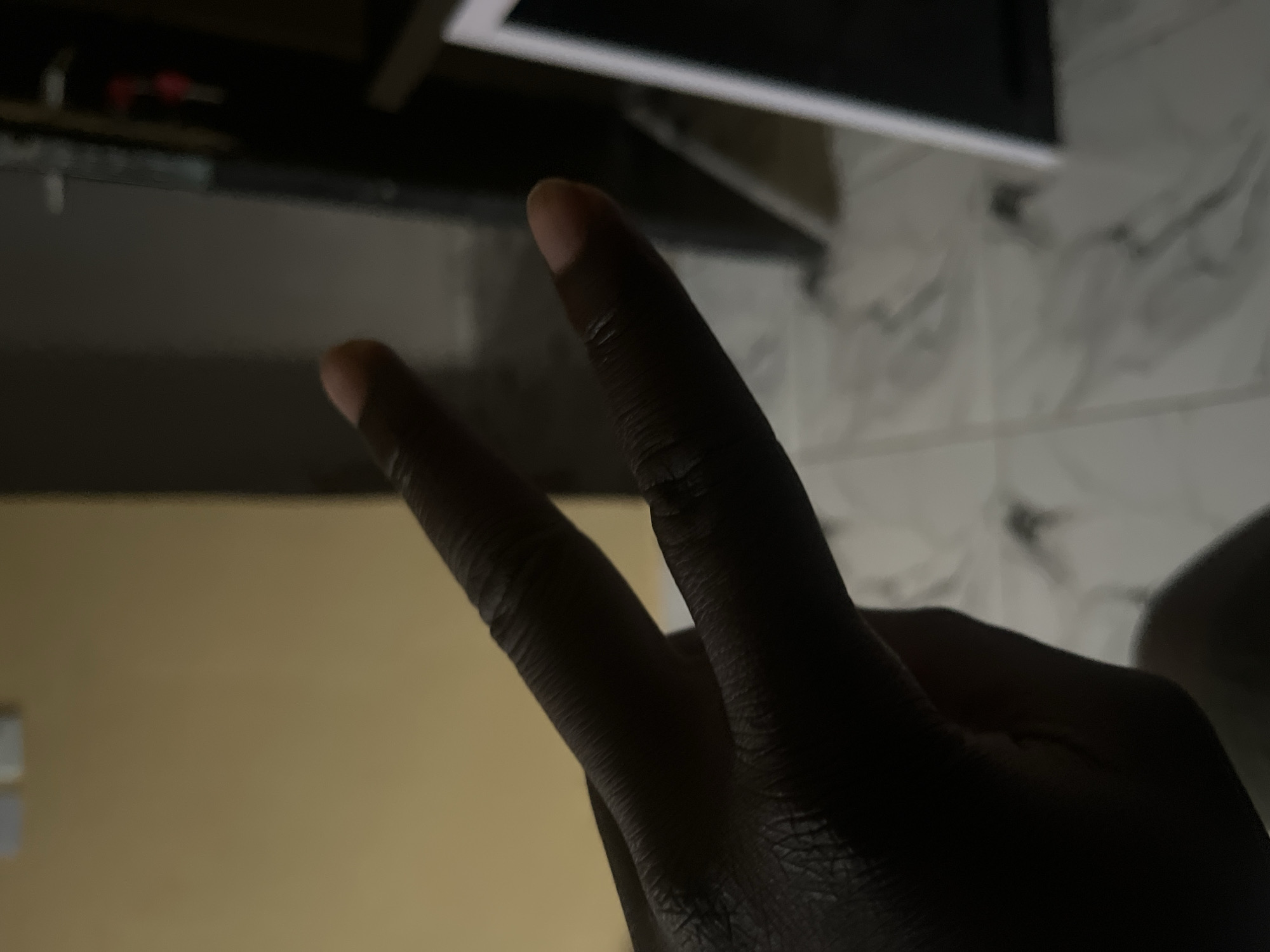 A hand is shown forming a peace sign, with the index finger and middle finger extended. The hand is dark-skinned and is positioned in the foreground, partially obscuring the background. The background consists of a yellow wall, a partially visible doorway with some objects, and a tiled floor with a marble pattern. The lighting in the image is dim, casting shadows. The scene is indoors, likely in a residential or commercial space. The location is Unknown, Unknown.