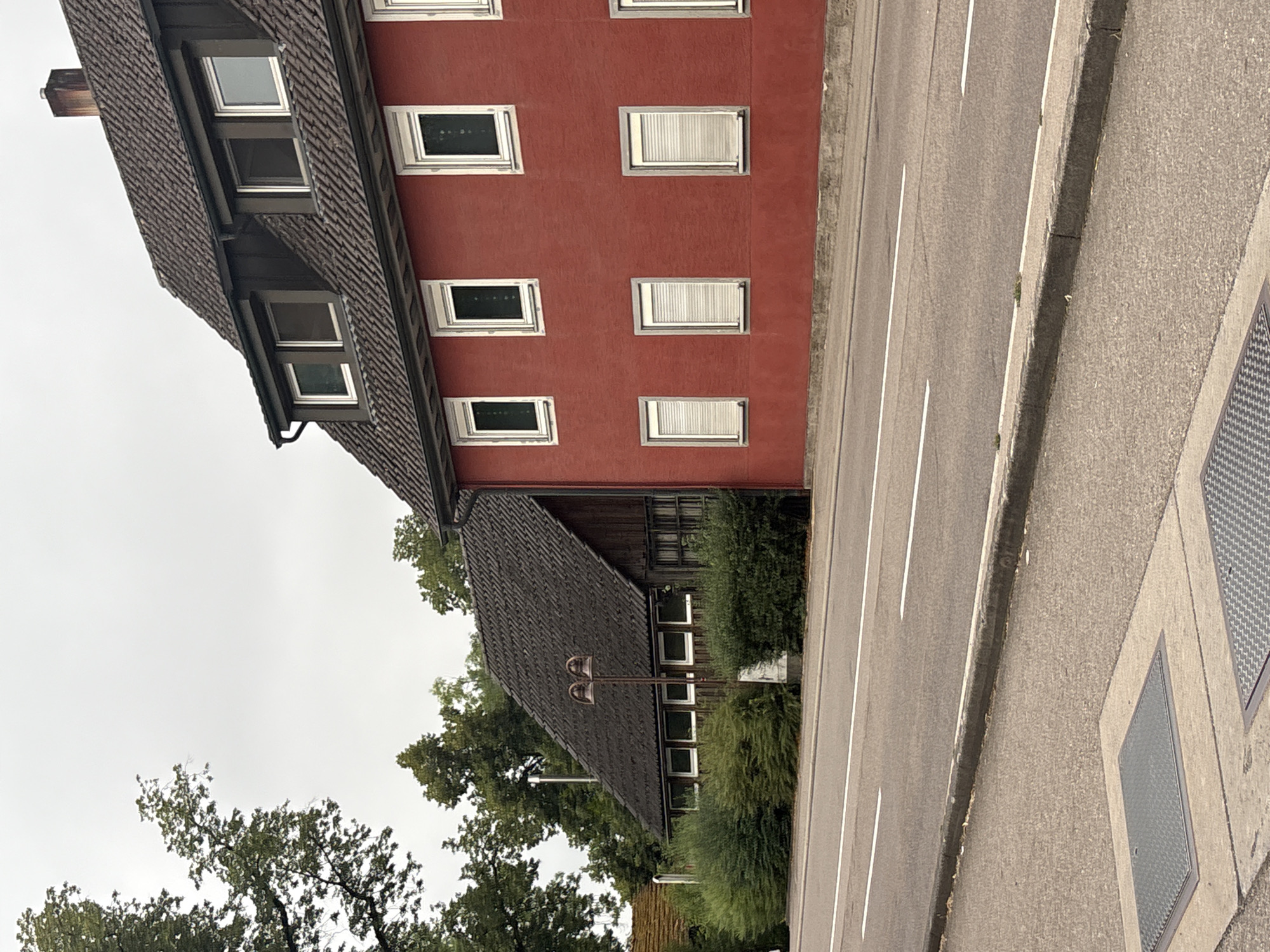 In Untermünkheim, Germany, a street scene unfolds. On the right, a red building with a dark tiled roof features multiple windows, some with closed shutters. Adjacent to it on the left is a lower, dark-tiled roof structure made of wood, also with several windows. Both buildings are bordered by green bushes. A paved road with white lane markings runs in front of the buildings, separated by a concrete curb. Two metal manhole covers are visible on the paved sidewalk area in the foreground. Trees are present in the background, and the sky is overcast.
