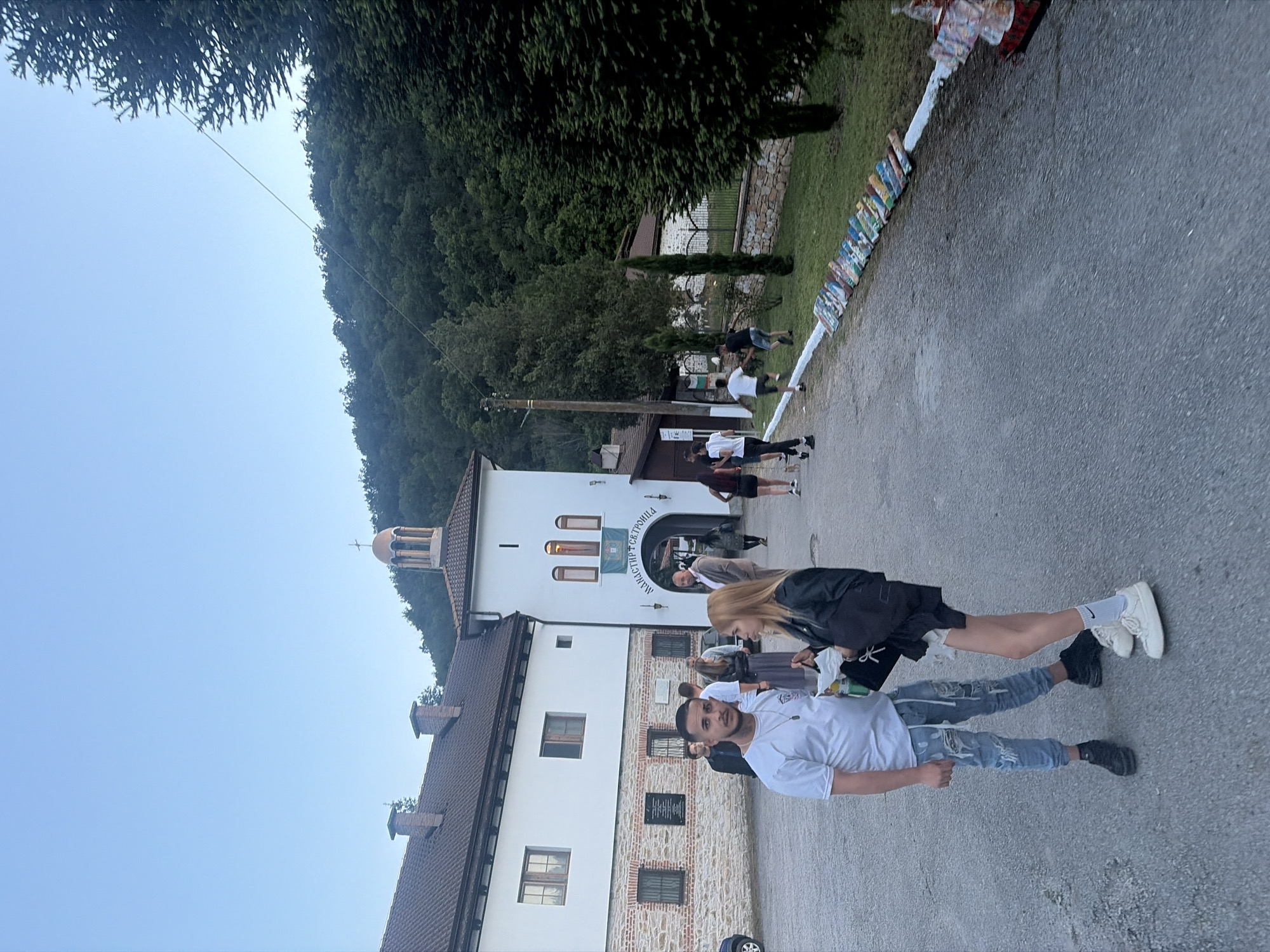 The image shows a group of people standing outside the Monastery of the Holy Trinity near the city of Mramor, Bulgaria. The monastery building, with its white facade, dark brown roof, and a small bell tower, is located on the left side of the frame. A group of people of varying ages are standing on the paved road in front of the monastery. A man in a white T-shirt and ripped jeans is standing closest to the camera, looking directly at it. A woman with blonde hair, wearing a black jacket and white sneakers, is standing next to him. Other people are standing further back, closer to the monastery entrance. The background includes a lush green forest covering the hills. On the right side of the road, there's a row of objects lined up, possibly paintings or other decorative items. The sky is clear and blue.