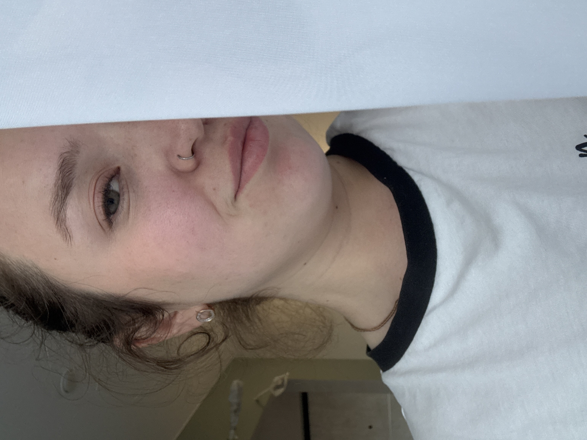 A close-up image shows a person with fair skin, light brown hair, and gray eyes. The person is wearing a white T-shirt with a black collar. The image is taken from a low angle. The person has a nose ring in their left nostril and a hoop earring in their left ear. Part of a white object, possibly a sheet or curtain, is visible above the person's head. The background appears to be a room interior with muted lighting. There is no information available to suggest this image was taken in Izhevsk, Russia, or any other specific location.