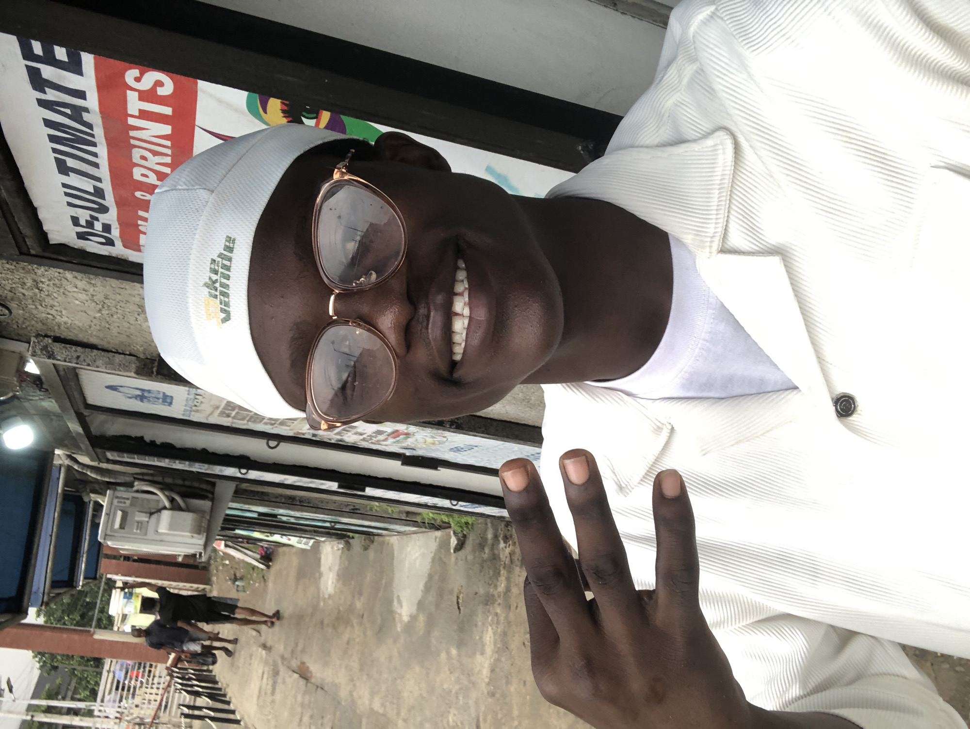 A young Black man, wearing a white t-shirt, white corduroy jacket, and white beanie with a yellow and green logo, smiles at the camera while holding up his left hand in a peace sign. He wears rose-gold rimmed glasses. The man is standing in front of a storefront in Ado, Nigeria. The storefront has a sign that reads "DE-ULTIMATE & PRINTS." To the left of the man, two other Black men are walking away from the camera down a paved walkway. One man is carrying a bucket on his head. Behind them, there is a brick wall, a blue awning, and an air conditioning unit mounted on the wall. The ground is cracked and uneven.