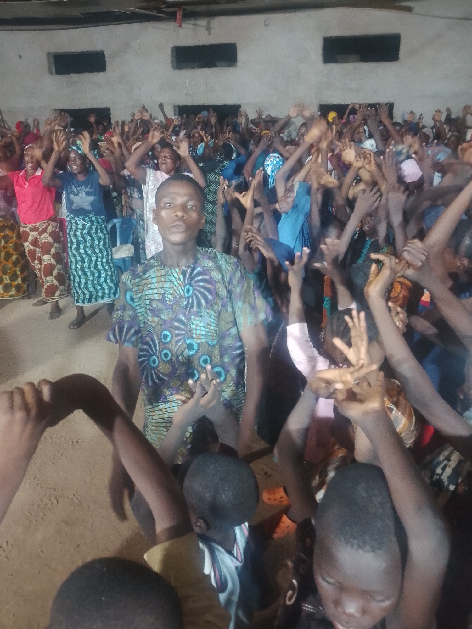In an indoor gathering, a young man in a patterned dashiki stands prominently in the foreground, facing the camera. He is surrounded by a large crowd of people, many of whom have their arms raised in the air, some with hands open and others in fists. The individuals in the crowd appear to be of various ages and genders, dressed in colorful clothing, including patterned skirts and headscarves. The environment suggests a community hall or church, with bare concrete walls and what appears to be a dirt floor. The lighting is dim, creating a somewhat grainy image. The overall atmosphere seems to be one of worship or celebration.