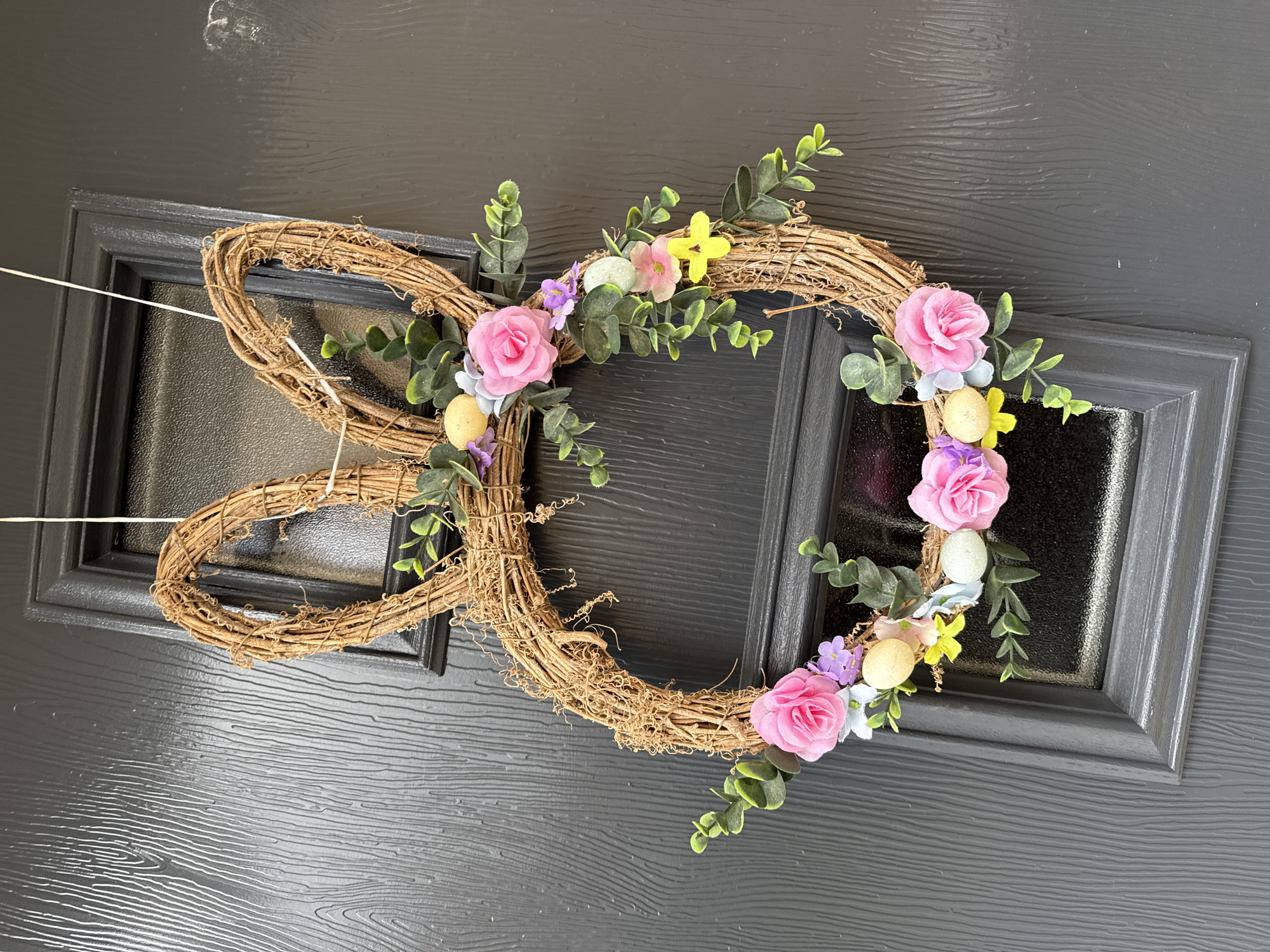 A close-up, eye-level shot presents a bunny-shaped wreath adorned with floral embellishments, suspended against a dark, textured door with inset windows. The wreath, constructed from woven twigs, forms the shape of a rabbit's head with two elongated ears. Pink, yellow, and purple flowers, alongside greenery and speckled eggs, decorate the wreath, primarily concentrated on one side. The dark door features a woodgrain pattern and inset windows framed in a matching dark color. Thin white strings suspend the wreath from the door. The location in the city of Great Yarmouth, United Kingdom is not evident from the image.