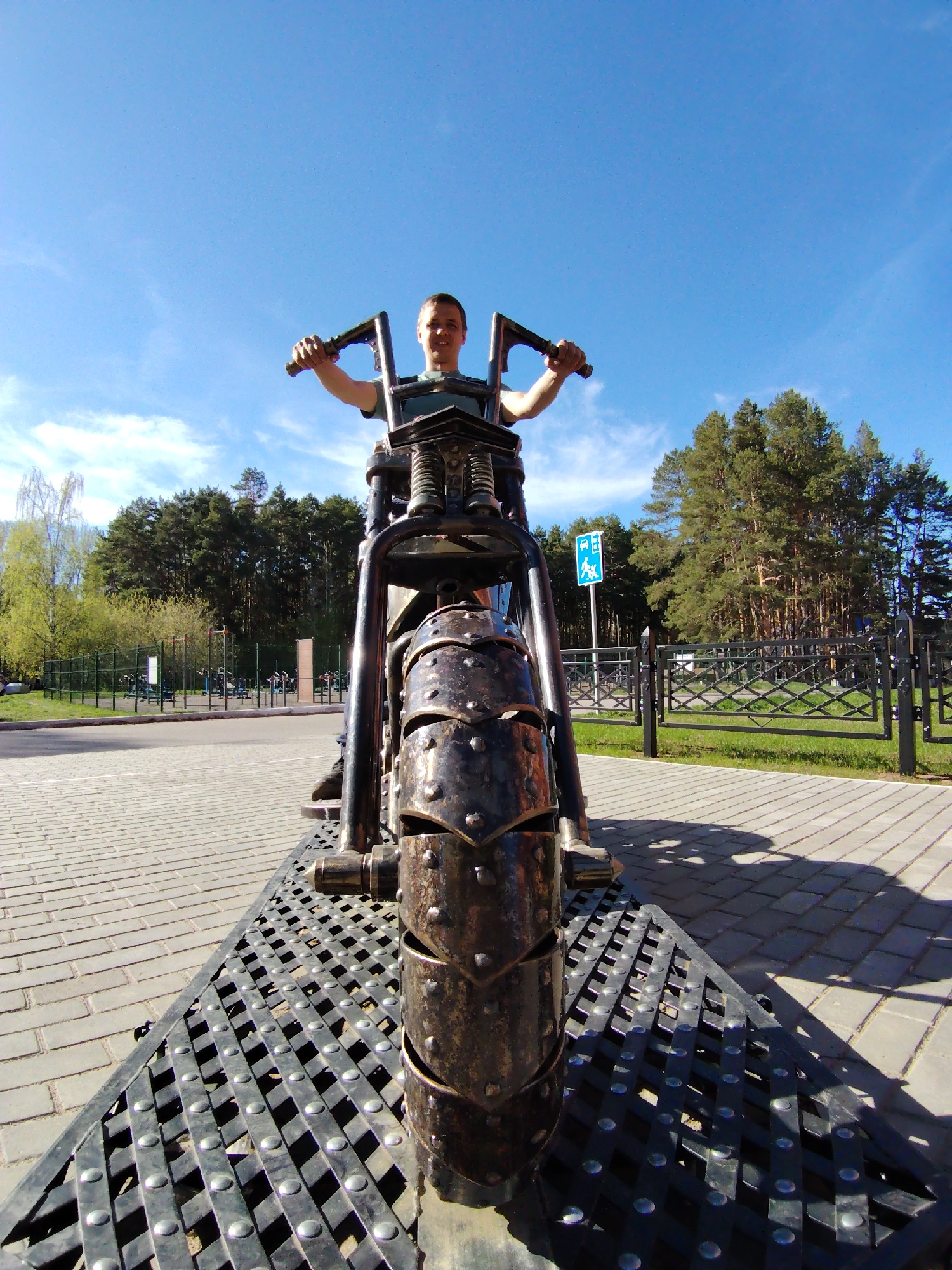 A man is posing on a metal motorcycle sculpture in Chaikovsky, Russia. The sculpture sits on a metal platform with a grate-like pattern. The ground is paved with gray bricks. A metal fence borders a grassy area with trees in the background. A blue sign with a white image is visible to the right. The sky is blue with scattered clouds.
