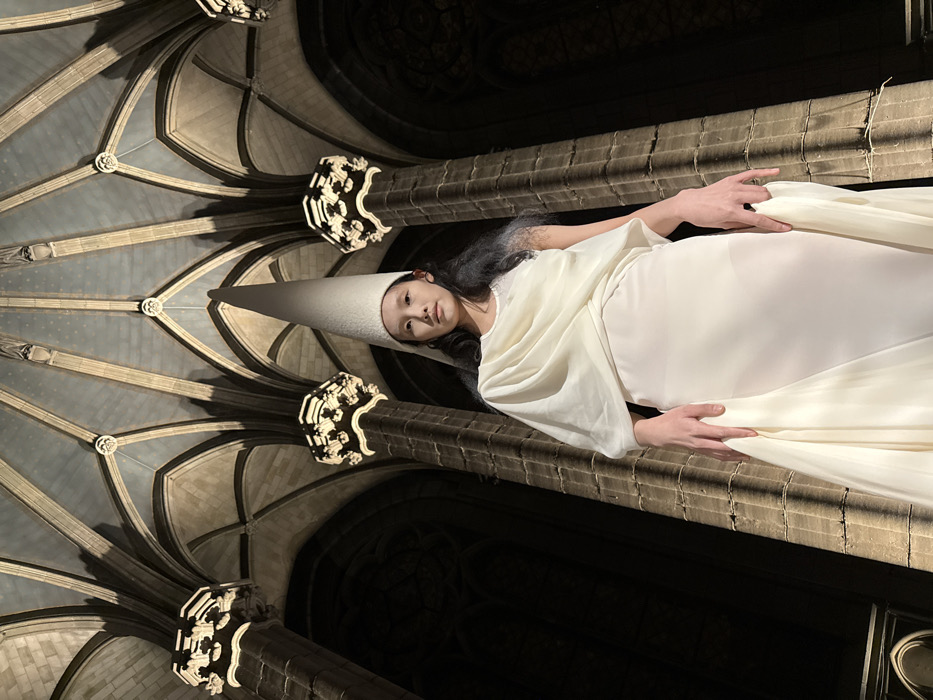 An individual in a white conical hat and flowing white gown lies amidst gothic architecture. The ribbed vault ceiling above is illuminated, contrasting with the dark columns and arches. The person's face is visible, and their hands rest on the stone structure. The image's orientation is rotated, with the person appearing to lie on their side.