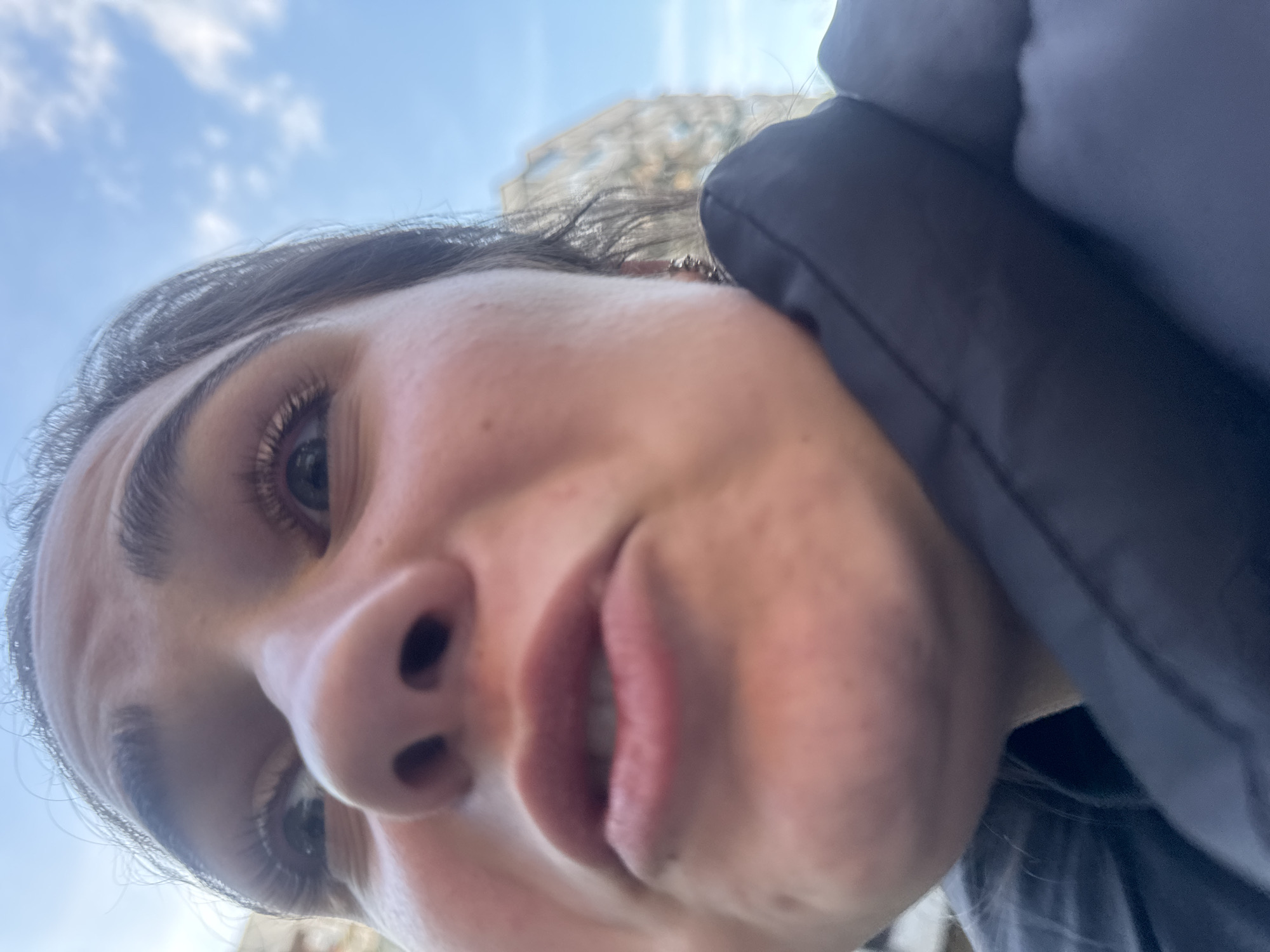 An extreme close-up, low-angle selfie captures a person looking directly into the camera from below. The subject is a female with dark hair, visible eyelashes, and a slightly open mouth revealing teeth. Her nose appears prominent due to the exaggerated perspective of the shot. She is wearing a dark-colored jacket or coat, the collar and shoulder of which are visible on the right side of the frame.
The background shows a light blue sky with scattered white clouds, indicating an outdoor setting during the daytime. A blurred, light-colored building or structure can be faintly seen in the upper right portion of the image. The lighting is bright and natural, illuminating the subject's face clearly. There are no visible texts or specific location cues beyond the suggestion of an outdoor environment. The person's expression is neutral to slightly relaxed, possibly a casual glance at the camera.