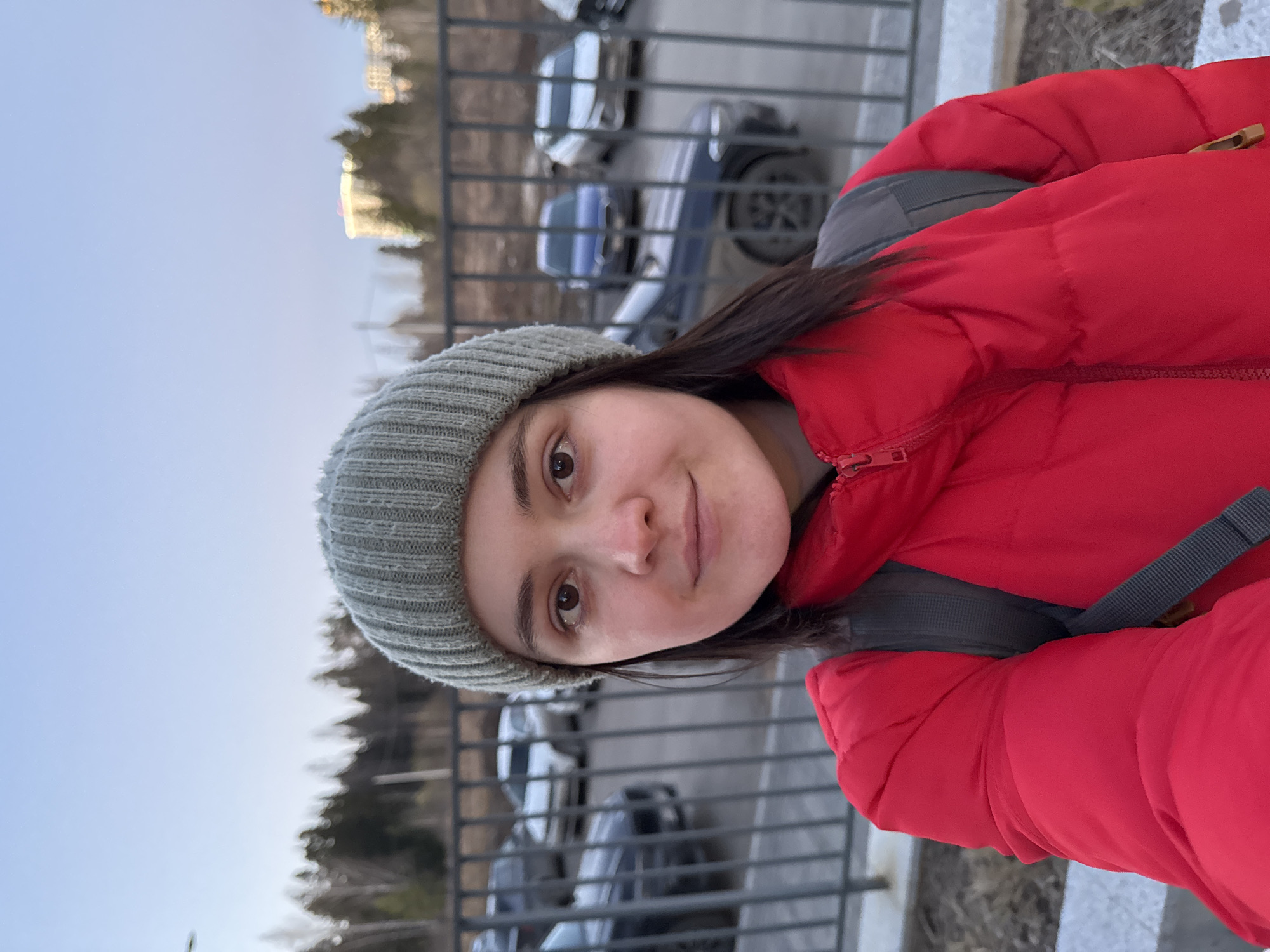 A woman in a red puffy jacket and gray knit hat stands in front of a metal fence. She is looking directly at the camera. Behind the fence is a parking lot with several cars. In the background, there are trees and a building. This image seems to be taken in Izhevsk, Russia.