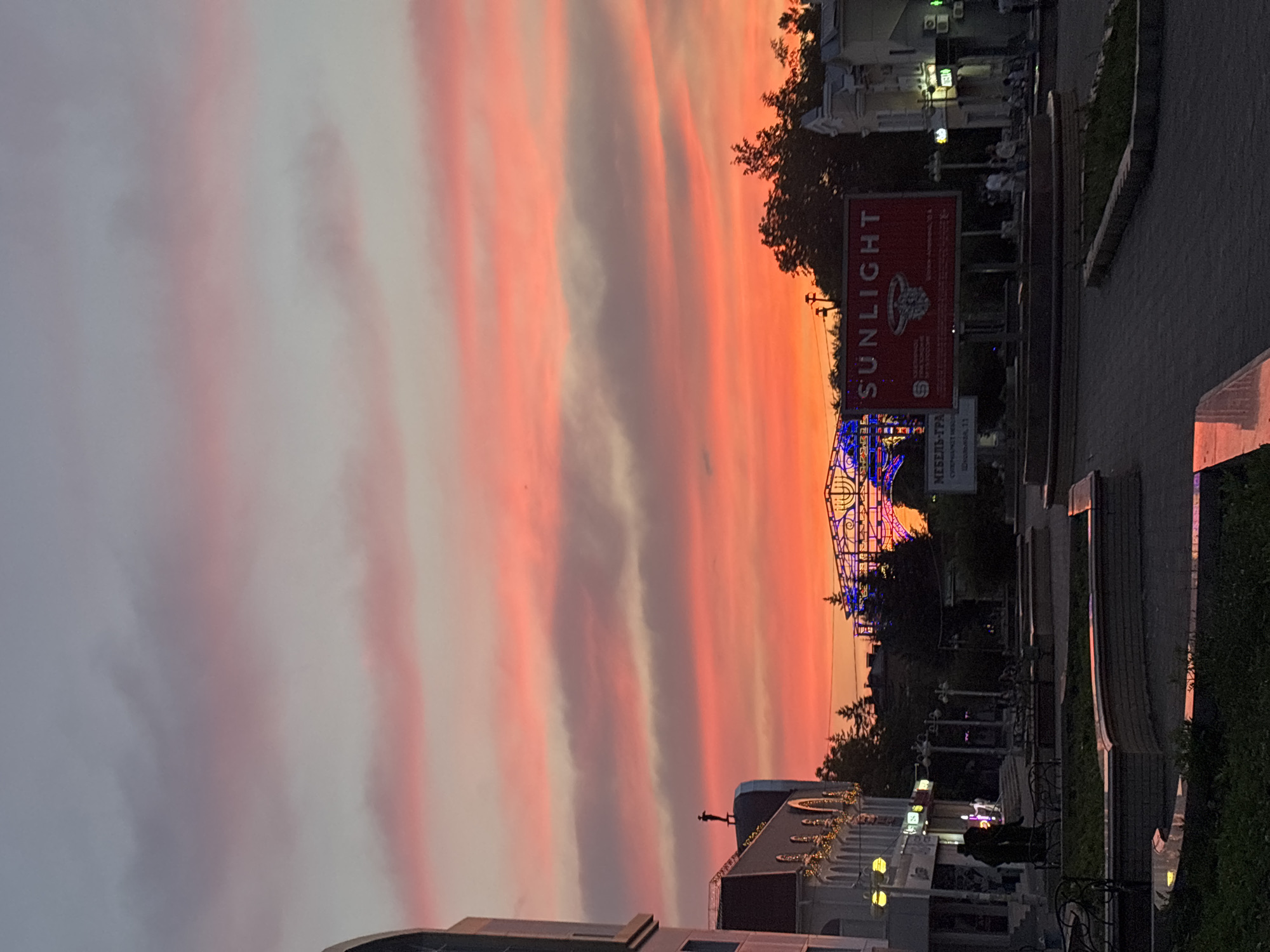 The image depicts a street scene in Birobidzhan, Russia, during a vibrant sunset. The sky is filled with dramatic clouds painted in hues of pink, orange, and red, suggesting a vivid twilight. In the foreground, paved walkways and decorative planters are visible, with benches suggesting a public seating area. Silhouetted figures, possibly people, are present near the walkways. Buildings line the street, some featuring illuminated windows and architectural details. A prominent red billboard with the word "SUNLIGHT" and an image of a ring is placed in the mid-ground. Behind the billboard, an arched structure adorned with blue and multi-colored lights is visible, potentially a decorative archway or part of an entertainment area. The overall atmosphere is one of a city transitioning from day to night, highlighted by the striking natural light display.