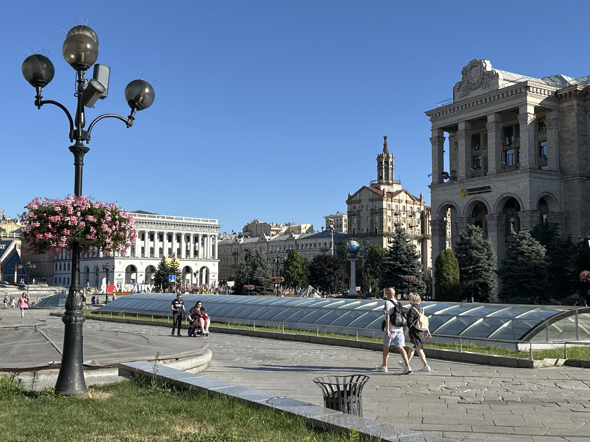 In Kyiv, Ukraine, on a clear, sunny day, a wide, paved plaza is depicted. Dominating the foreground is a black lamppost with three globe-shaped lights and a hanging basket of pink flowers. To the right, a path leads towards a large, modern building with a grand, classical facade featuring numerous columns and arches. A yellow logo and text "УКРПОШТА" are visible on the building, indicating it is likely a post office. In the mid-ground, a group of people are present; one individual is in a wheelchair, accompanied by another person. Further back, more individuals stroll across the plaza, and several trees and smaller buildings are visible. A long, transparent, sloped roof structure runs across the mid-ground, possibly covering an underground passage or a display area. In the background, more multi-story buildings form the urban landscape. The sky is a vibrant, cloudless blue.