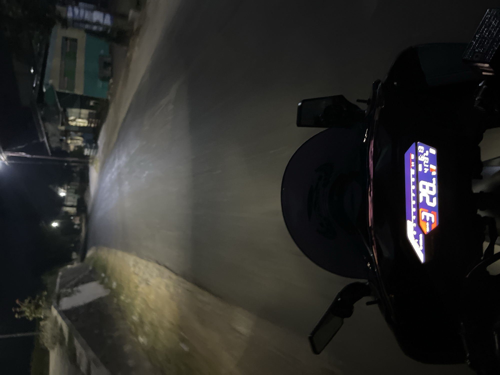 The image is a first-person perspective of someone riding a motorcycle at night on a paved road in Karangrandu, Indonesia. The road is illuminated by the motorcycle's headlights, which cast a strong beam forward, creating a tunnel of light. On either side of the road are dimly lit areas with vegetation and what appear to be simple structures, suggesting a rural or semi-urban setting. One side of the road has a low wall and some scattered debris, while the other side shows buildings, one of which has a teal and white facade and appears to be a shop or residence. The motorcycle's digital dashboard is visible in the foreground, displaying glowing orange numbers "3 28" and other symbols. The overall atmosphere is dark and quiet, characteristic of a nighttime scene in a residential or less developed area.