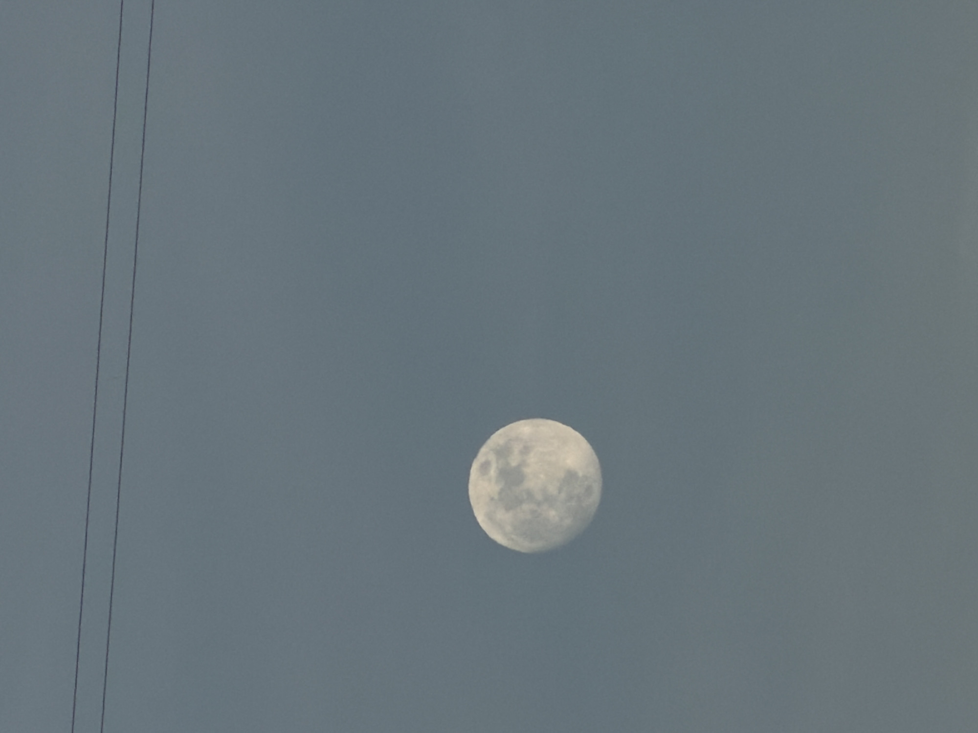 A medium shot captures the moon against a gray-blue sky, with two power lines visible on the left. The location is undetermined, and there's no indication of the city of Chaikovsky, Russia, or any people, actions, or interactions in the frame.