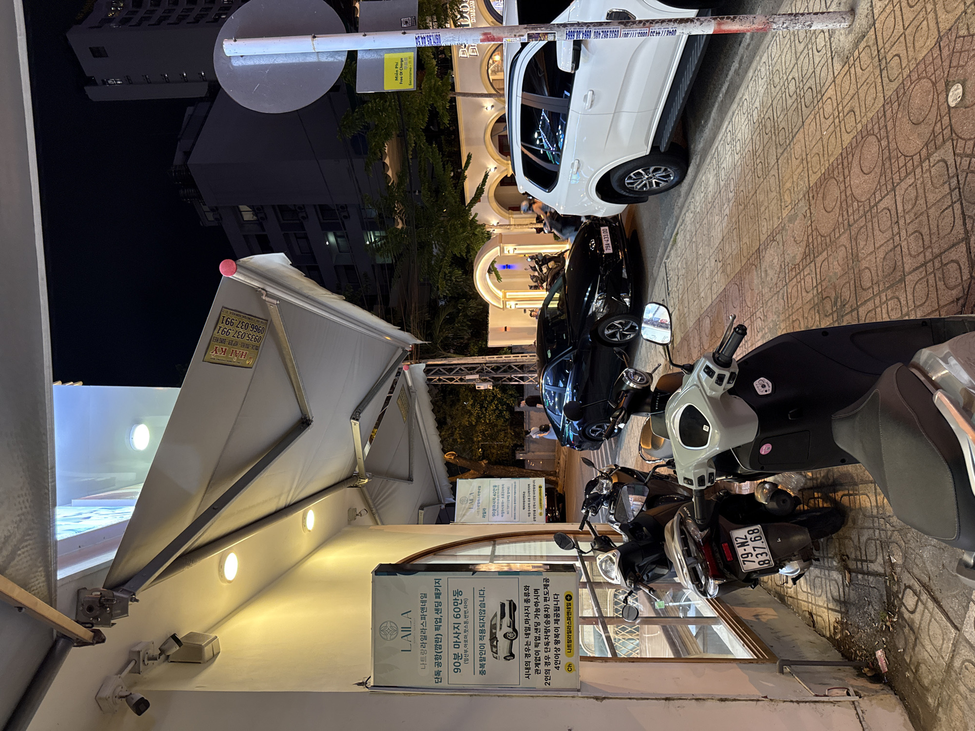The image depicts a street scene at night in Nha Trang, Vietnam. A white SUV is parked on the right side of the frame, with a black sedan parked behind it. Several motorbikes are parked along the sidewalk on the left. A sign on a building reads "LAILA" and appears to be advertising services. The sidewalk is paved with patterned tiles, and there are trees and buildings in the background. The lighting suggests it is nighttime, with artificial lights illuminating the storefronts and street.