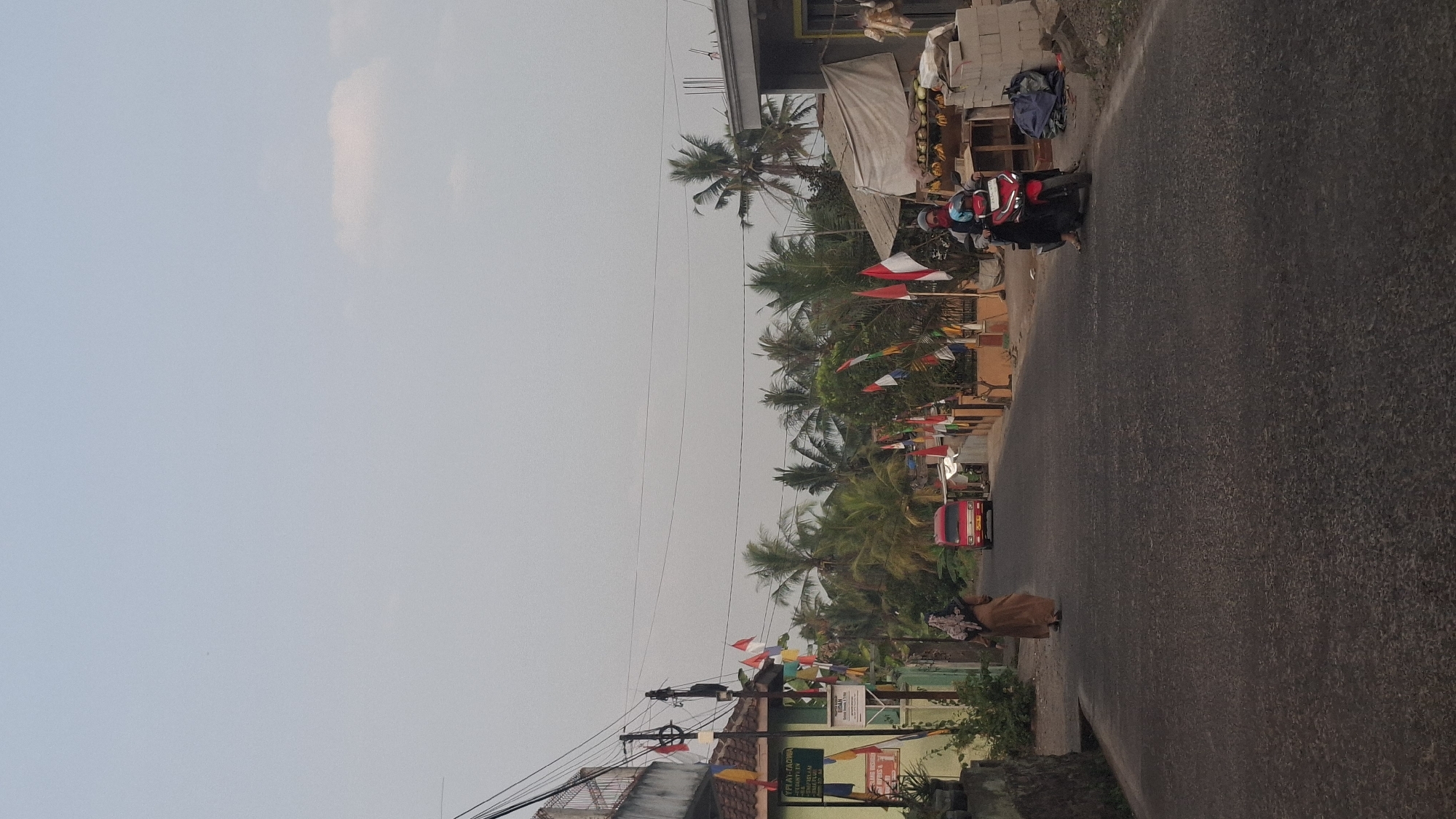 The image shows a street scene in Cibinong Hilir, Indonesia. The road is paved with asphalt and lined with trees, mostly palm trees. On the left side of the road, there is a building with a green facade and a sign. Small Indonesian flags are hung along the street and on poles. A red car is driving away from the camera down the road. Further down the road, on the right side, there is a market stall with fruits. Two people are visible: a person in a long brown garment and headscarf walking away from the camera on the left side of the road, and another person on a red motorcycle wearing a helmet and a blue headscarf, also moving away from the camera on the right side of the road. The sky is overcast.