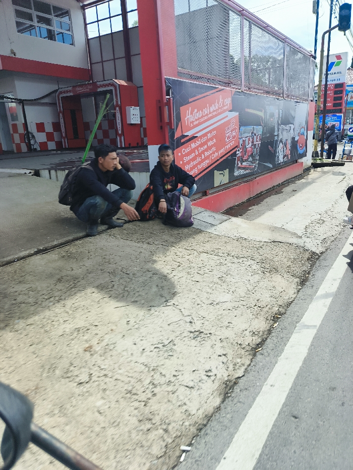 The scene takes place in Ciwidey, Indonesia. Two young men are seated on the sidewalk in front of a car wash establishment. The men are wearing casual clothing. They are sitting with their backs against the building. One man has a black backpack on and the other has a dark green backpack on. Both men have their legs crossed. One man has his right hand on his lap and the other man is holding his left hand in front of him. The car wash has a large sign that says "Hotline car wash & care" in yellow letters. The sign also lists the car wash services offered in Indonesian. There is a white line painted on the edge of the road to the right of the sidewalk. The road is paved. The building is red and white. The sidewalk is made of gray concrete. There is a small green plant growing in the cracks of the sidewalk. The building has a large window on the left side and a small window on the right side. There is a green pole sticking out of the building on the left side. There is a grate on the left side of the sidewalk. There is a large metal grate on the right side of the building. There is a large advertisement for the car wash on the right side of the building. The sign shows a drawing of a large car being washed. There are no cars or vehicles visible in the scene. There is one cigarette butt on the sidewalk near the white line.