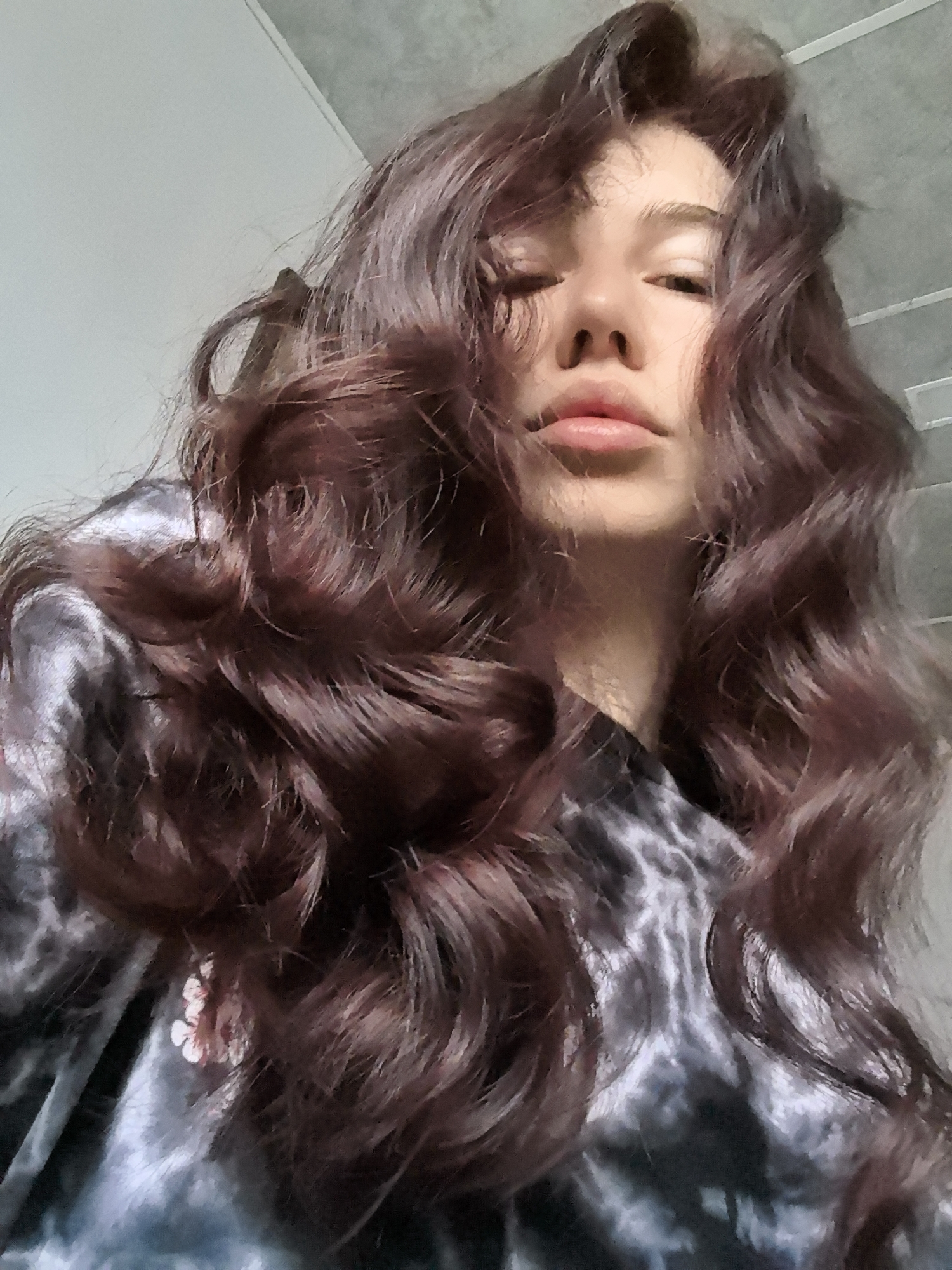 An upward-angled close-up photograph depicts a single individual, presumably female, in an indoor setting within Stara Zagora, Bulgaria. The subject's head and upper torso are visible, with long, wavy, dark reddish-brown hair cascading around the face and over the shoulders. The individual's eyes are mostly closed, and the mouth is slightly parted with full lips. The facial features include a prominent nose and smooth skin. The subject is wearing a dark garment with a black, grey, and white mottled or tie-dye pattern. A small, light-colored floral or abstract design is present on the left shoulder area of the clothing. The background consists of a light-colored (off-white/light grey) wall on the upper left and a dark grey, textured ceiling with faint linear patterns occupying the upper right and central top portions of the frame. The lighting appears soft and diffused, illuminating the subject from above.