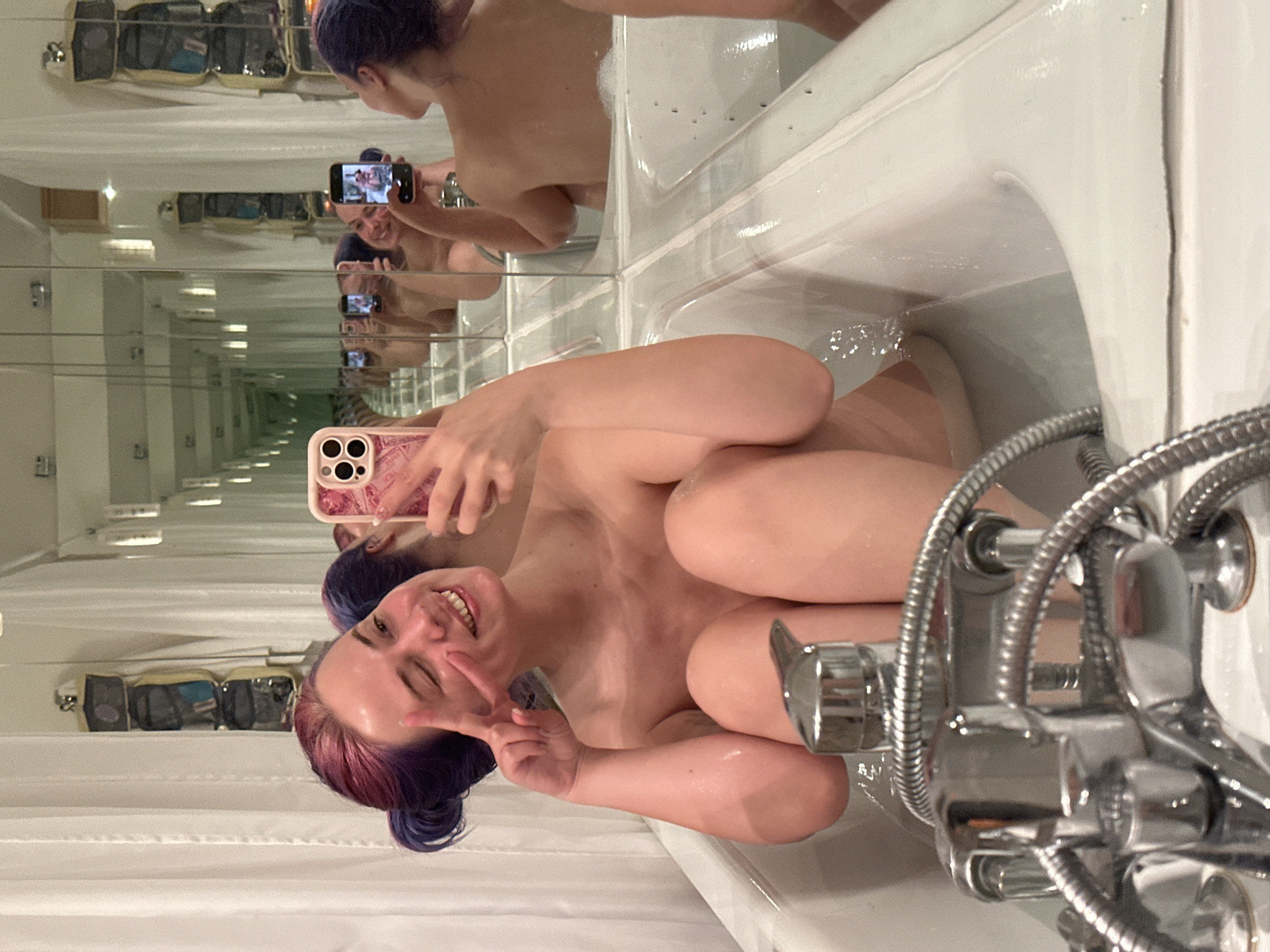 A woman with dyed purple and red hair is taking a selfie in a bathtub filled with water. She is making a peace sign with her right hand and smiling. The bathtub is white, and there is a chrome faucet and shower hose to her right. Behind her are mirrored walls, creating an infinity effect of reflections. A toiletry bag is visible on a shelf in one of the mirrored reflections. The location is unknown and has not been disclosed.