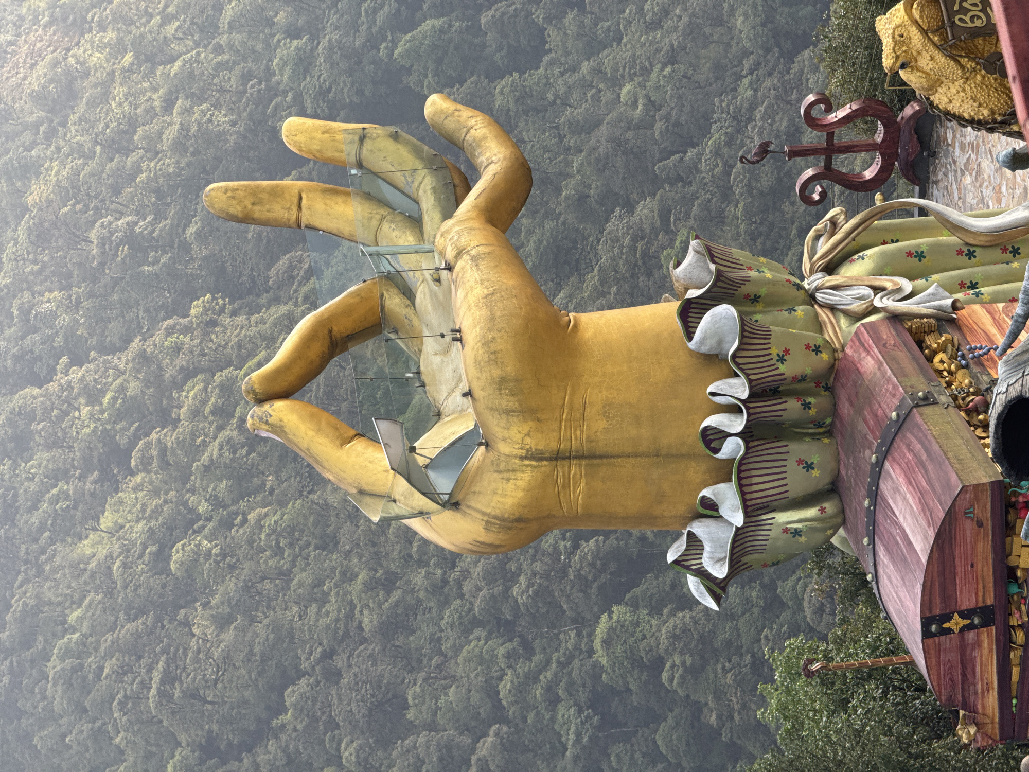 A large, yellow, sculpted hand protrudes over a green, dense forest landscape. The hand has glass panels between the fingers, supported by metal brackets. The base of the hand is adorned with a frilly, colorful cuff. To the right, there's a dark-red, barrel-like object with metal bands and decorative elements. The setting is the city of Binh Lu, Vietnam.