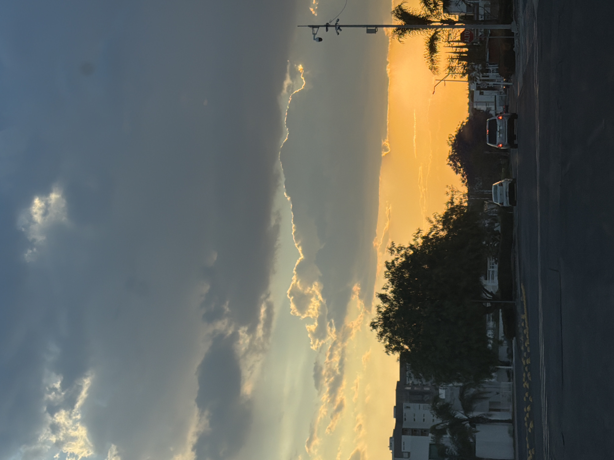 The image captures a sunset scene in an urban environment. A street with a dark asphalt road runs vertically along the right side. Buildings are visible on the right. Two vehicles are driving on the road. Overhead, a dark, cloudy sky contrasts sharply with a bright, orange and yellow sunset. Palm trees and a surveillance camera attached to a pole can be seen.
