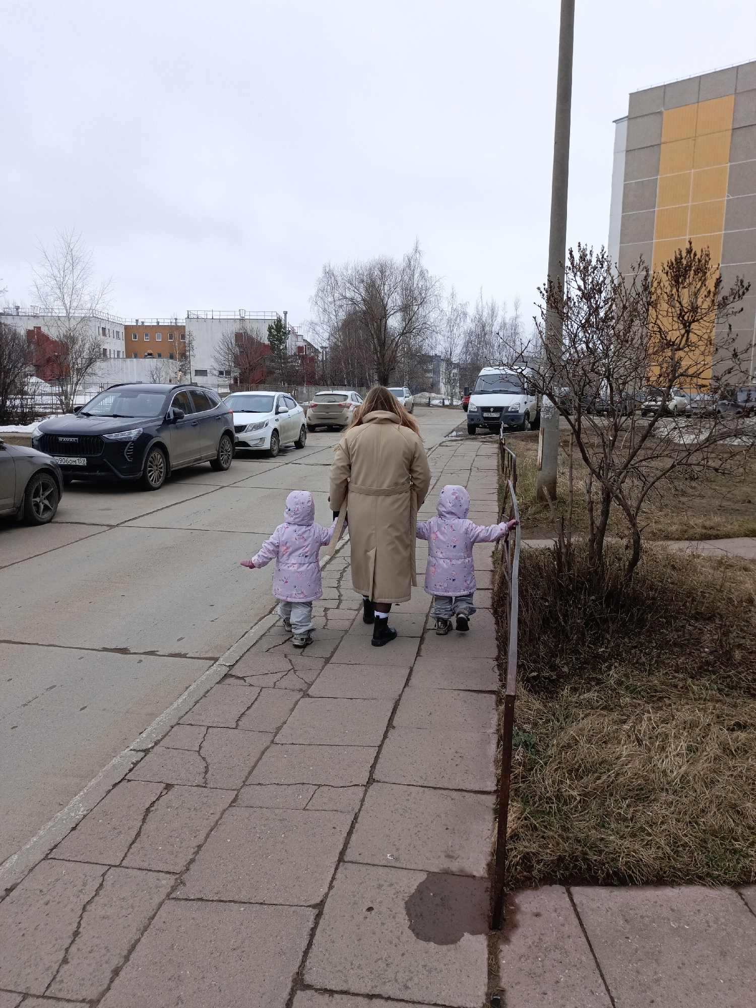 The image shows a woman and two young children walking on a sidewalk. They are walking away from the camera. The woman is wearing a beige coat and black shoes. The children are wearing matching purple hooded jackets and grey pants. To the left of the sidewalk is a road with several parked cars. In the background, there are apartment buildings and trees. The sky is overcast. This scene is likely located in the city of Chaikovsky, Russia based on the license plate number visible on the black car.