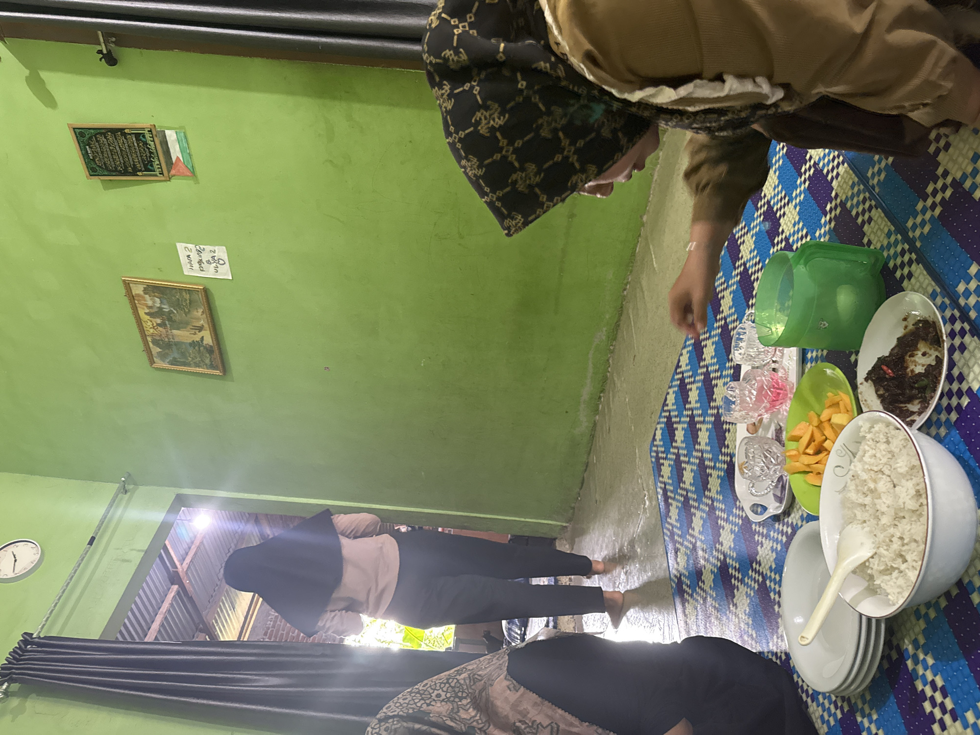This is a medium shot, taken from a low angle, of a group of people eating a meal on the floor of a room in Babahjurong, Indonesia. The room has green walls and a patterned mat on the floor. In the foreground, there is a plate of rice, a plate of French fries, and a plate of some kind of dark, saucy food. There is also a green pitcher and several glasses on the mat. Two women are visible in the foreground, both wearing hijabs. One woman, in the foreground right, is wearing a brown hijab and a brown top. The other woman, on the far left, is partially visible, wearing a dark hijab and a patterned top. In the background, a third woman in a black hijab and a light brown top is walking away from the camera towards an open doorway. The doorway leads outside to what appears to be a corrugated metal roof structure. On the wall to the right, there is a framed picture and a framed piece of calligraphy with the Palestinian flag underneath. A clock is visible on the wall to the left. The lighting is dim, with light coming from the doorway.