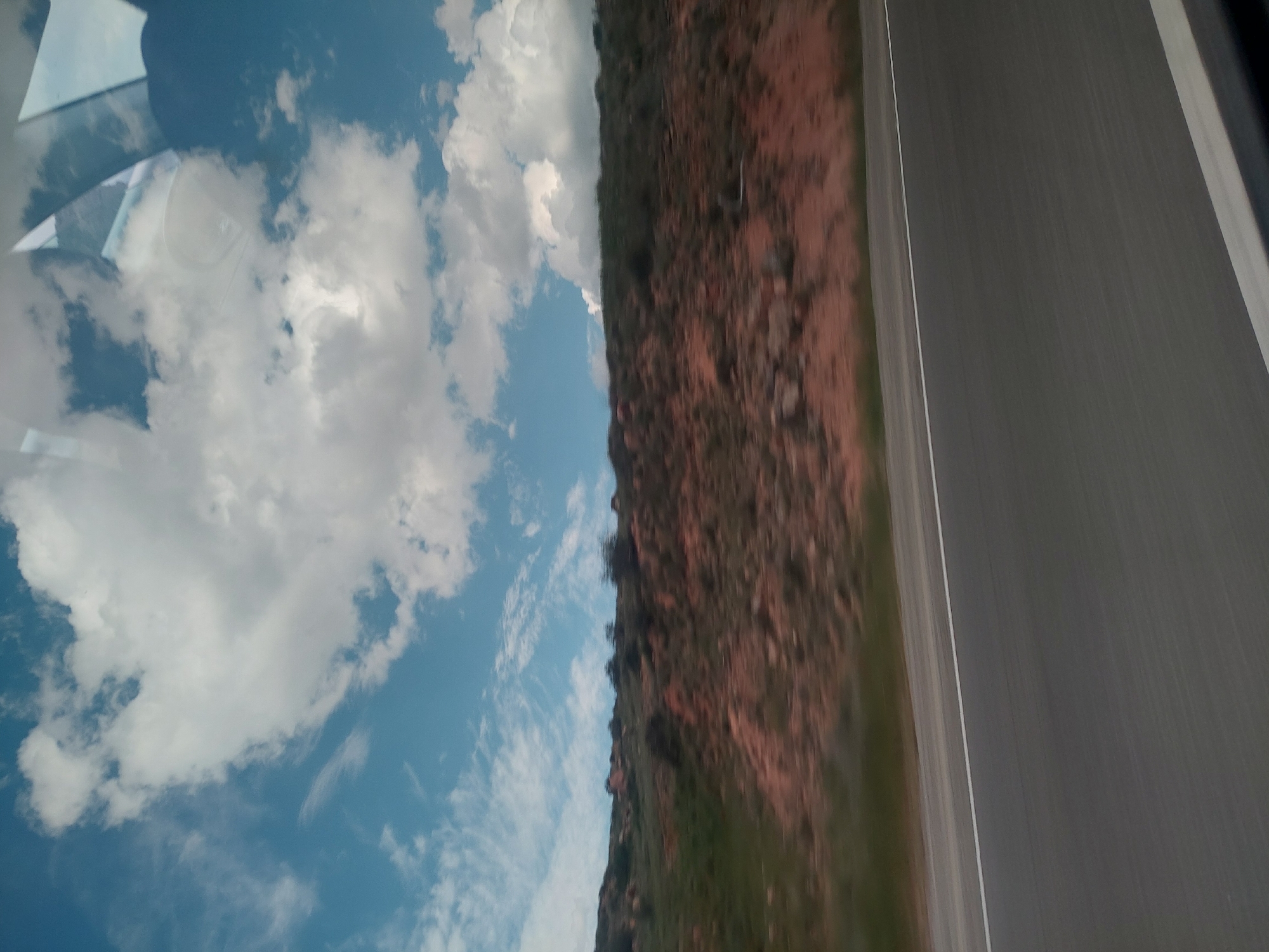 The image captures a landscape scene, likely viewed from a moving vehicle, as suggested by the motion blur on the road and reflections on a window pane. The photograph appears to have been taken with the camera rotated, presenting the road and land vertically, and the sky horizontally.
When re-oriented, the scene depicts a wide, grey asphalt road across the bottom of the frame, featuring a white solid line along its edge. The blurred appearance of the road indicates movement. Adjacent to the road, a rugged, reddish-brown hillside or mountainous terrain dominates the mid-ground, partially covered with patches of green vegetation and scattered shrubs. Above this natural landscape, a bright blue sky is filled with a dynamic array of white clouds, ranging from large, fluffy cumulus formations to more delicate, wispy streaks.
In the upper left corner, reflections are visible on what appears to be a vehicle window, showing indistinct shapes possibly from the car's interior or blurred external elements. The clear blue sky and strong illumination suggest it is daytime, under fair weather conditions. The composition evokes a sense of travel through an open, natural environment. No discernible text is visible within the image. The provided location context is Tigranashen, Armenia.