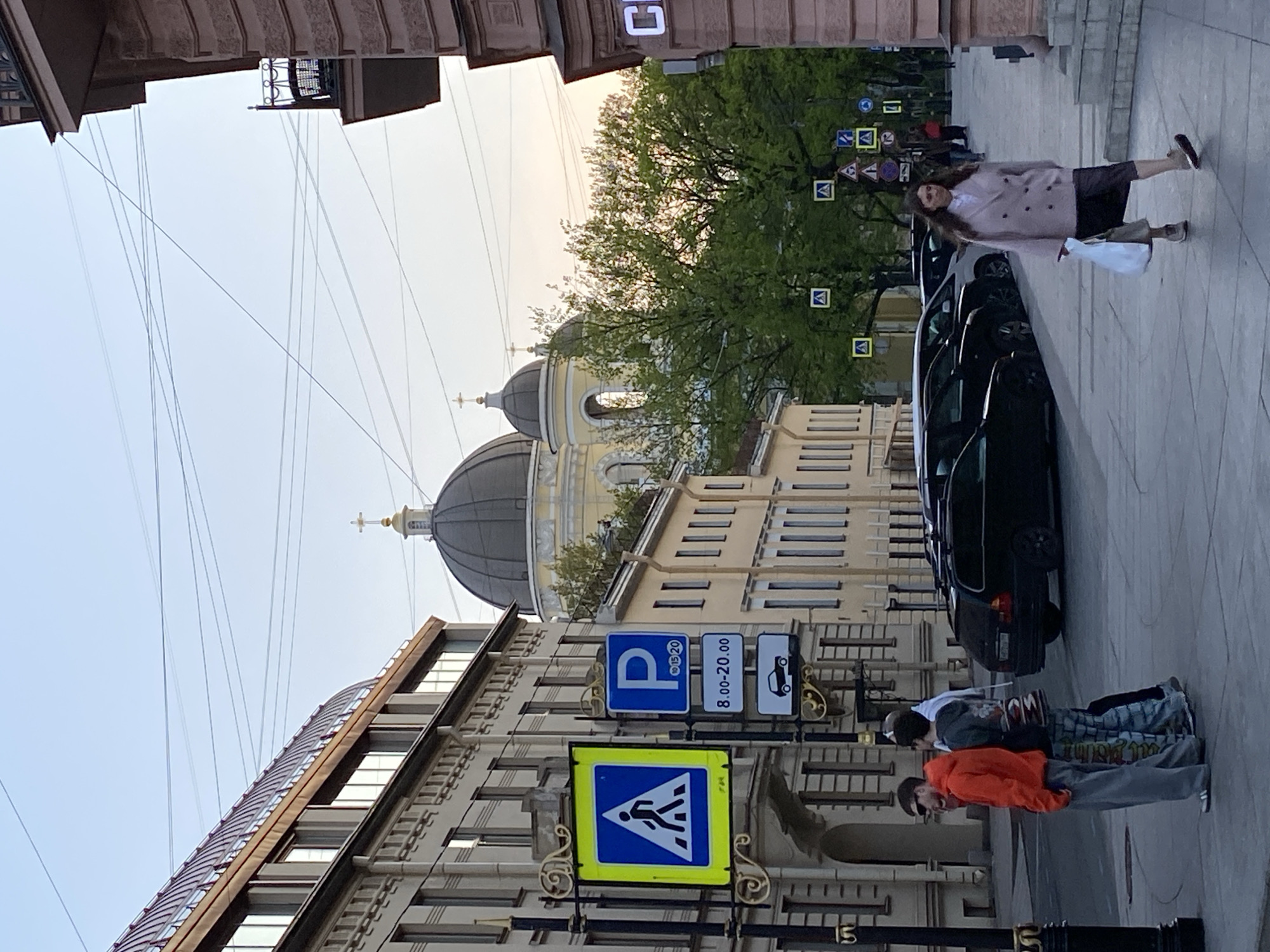 The image shows a street scene in Saint Petersburg, Russia. Dominating the background are the domes of Saint Catherine's Armenian Church, a prominent architectural landmark. A yellow building is visible behind the church.
The streetscape includes pedestrians, parked cars, and street signs. A person with long hair is walking on the right side of the frame. There are several parked cars, and a group of two men stands near a crosswalk sign. The street sign features a pedestrian crossing symbol, indicating a designated area for pedestrians to cross the street. Above it are parking regulation signs.
The buildings lining the street feature historical architectural details. Overhead, electrical wires run between the buildings. The sky is overcast, suggesting twilight or a cloudy day.