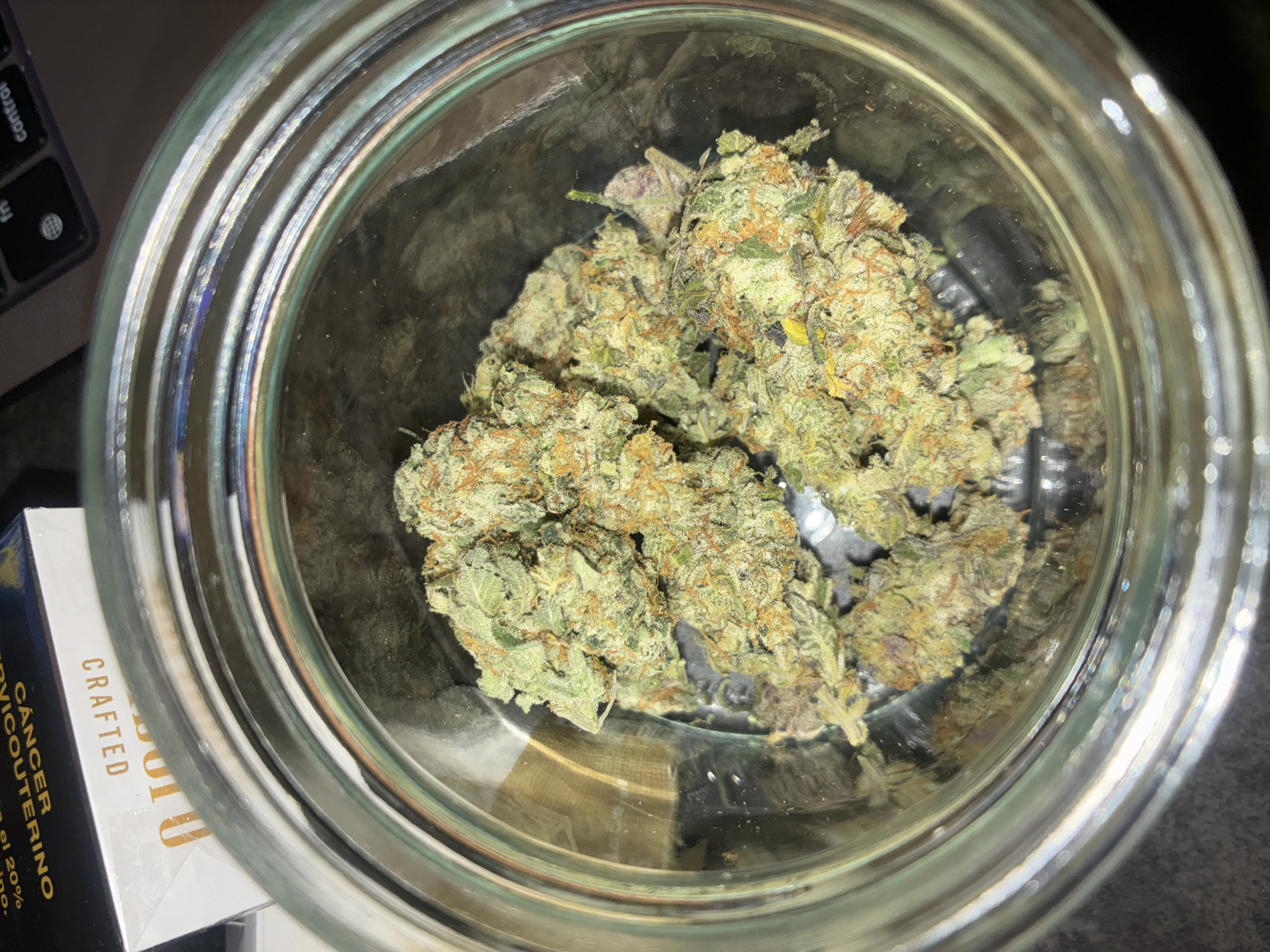A close-up, high-angle shot depicts a clear glass jar filled with cannabis buds. The buds are predominantly green with orange pistils and a light dusting of trichomes. The jar is partially positioned over a box with the text "CANCER CERVICOUTERINO" printed on it. Parts of a laptop keyboard are also visible in the top left corner of the image. The location details are not discernible from the image. Therefore, information about Huixquilucan de Degollado, Mexico, is not possible to extract.