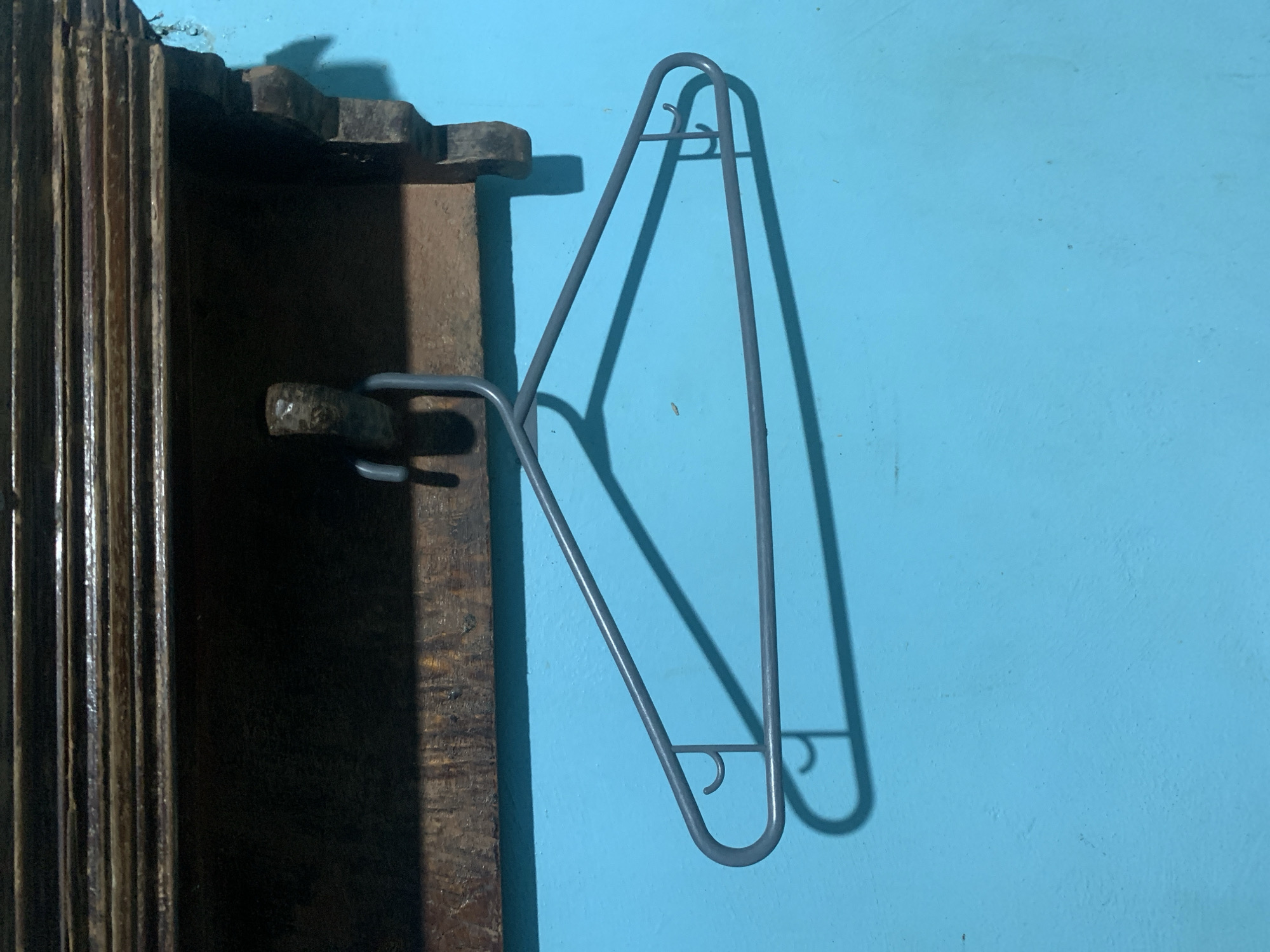 The image displays a static indoor or sheltered scene, primarily featuring a grey metal clothes hanger set against a vibrant blue wall. The hanger, designed with multiple bars and integrated hooks, casts a distinct, sharp shadow on the blue surface, indicating a strong light source originating from the left.
To the left of the frame, a dark, rustic wooden structure occupies about a quarter of the image. This structure, possibly part of a door frame, window, or furniture, exhibits vertical grooves or slats and has a weathered, dark brown appearance. A small, rough-textured, dark element, perhaps a hook or a projection, extends from this wooden structure towards the hanger.
The blue wall serves as the main background, showing minor surface irregularities consistent with painted concrete or stucco. There are no people visible, and no specific activity or event is taking place; the hanger appears to be simply mounted or placed on the wall. The defined shadows suggest strong illumination, possibly from natural daylight entering a room or from an artificial light source. The overall aesthetic, with its functional objects and simple background, aligns with a typical domestic setting in a location like Santa Ana, Venezuela. No visible text is present in the image.