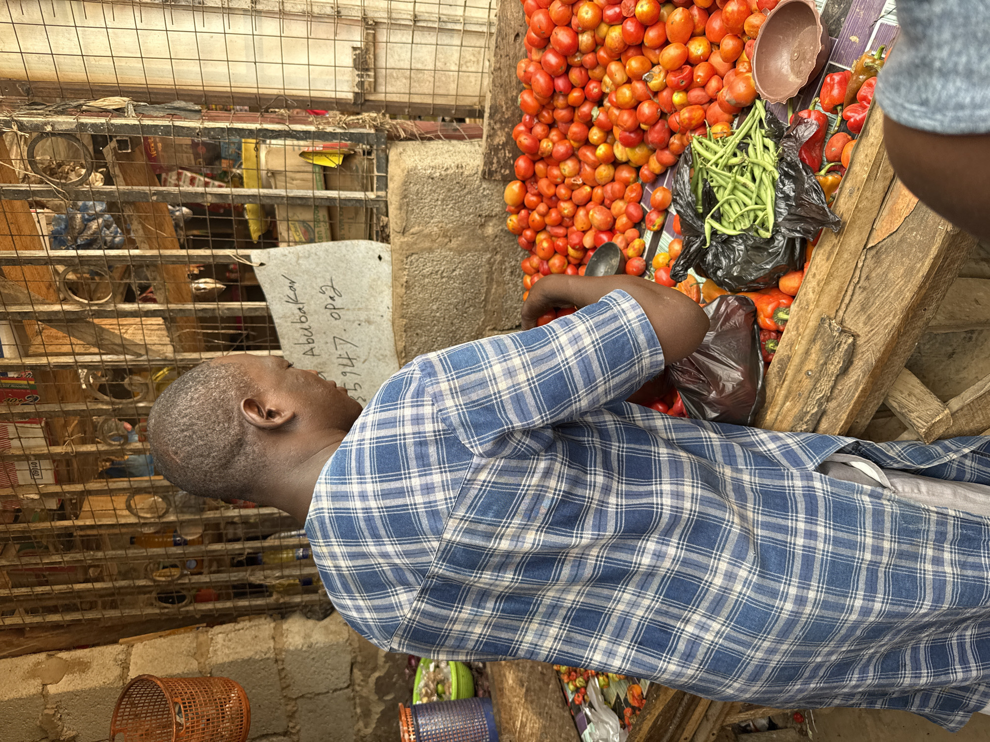 An adult man wearing a blue and white plaid shirt is standing at a market stall in Abuja, Nigeria. He is selecting tomatoes from a large pile of ripe, red tomatoes and placing them into a black plastic bag. The stall also contains green beans, red and orange bell peppers, and a brown bowl. The man is positioned near a wire cage filled with various items. Next to the cage, there is a piece of paper with handwriting on it.