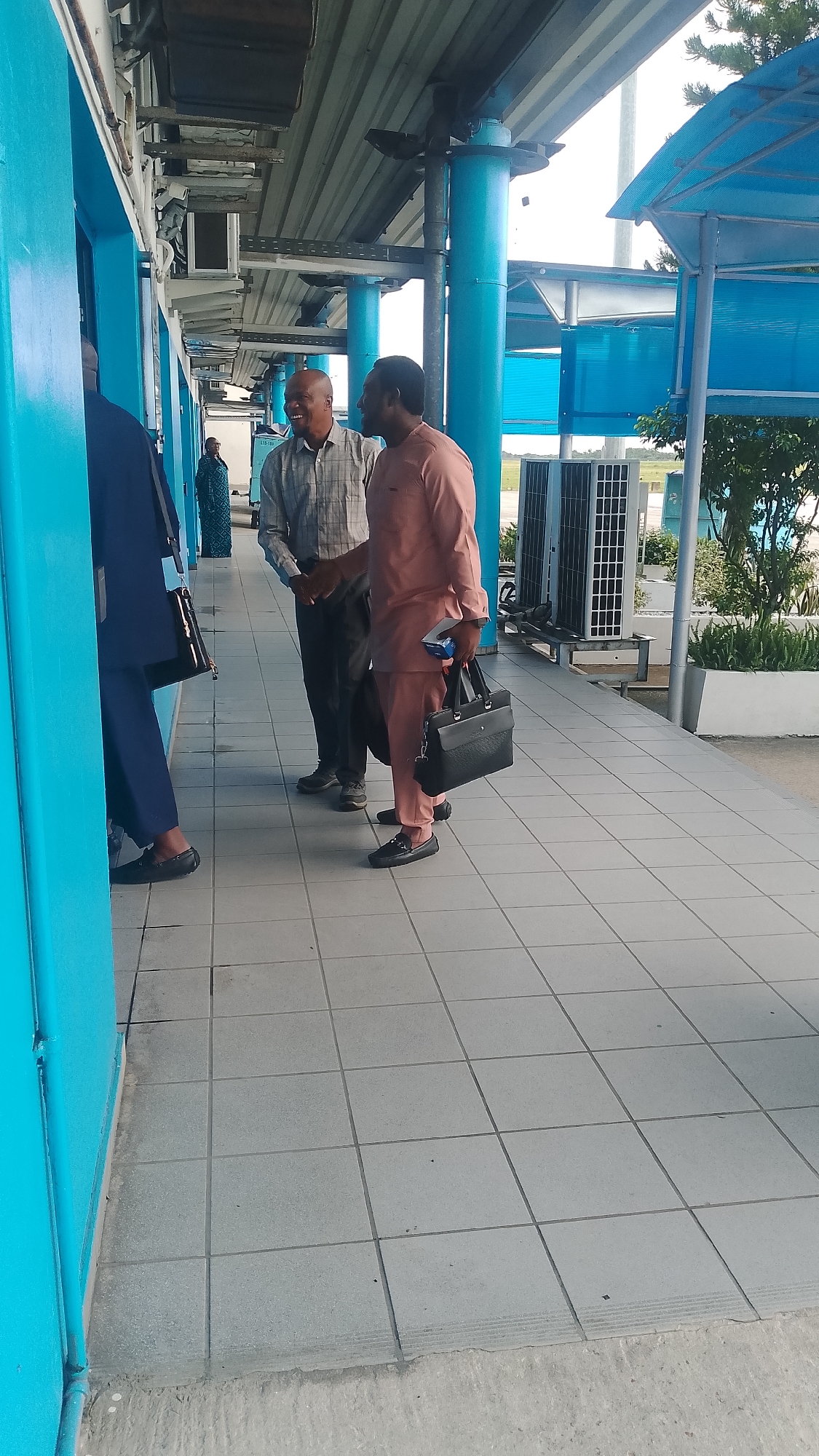 Three men are standing on a tiled walkway outside a building. The building has bright blue walls and a metal roof supported by blue pillars. The man on the left is wearing a dark blue suit and is partially obscured by the wall. The man in the middle is wearing a plaid shirt and dark pants, and is shaking hands with the man on the right. The man on the right is wearing a peach-colored traditional outfit and is holding a black briefcase. Two large air conditioning units are visible in the background. The scene appears to be outdoors, possibly in a public area or an airport, given the AC units and the walkway. The lighting suggests it is daytime, and the weather appears to be fair. The men's body language, particularly the handshake and smiles, suggests a positive interaction.