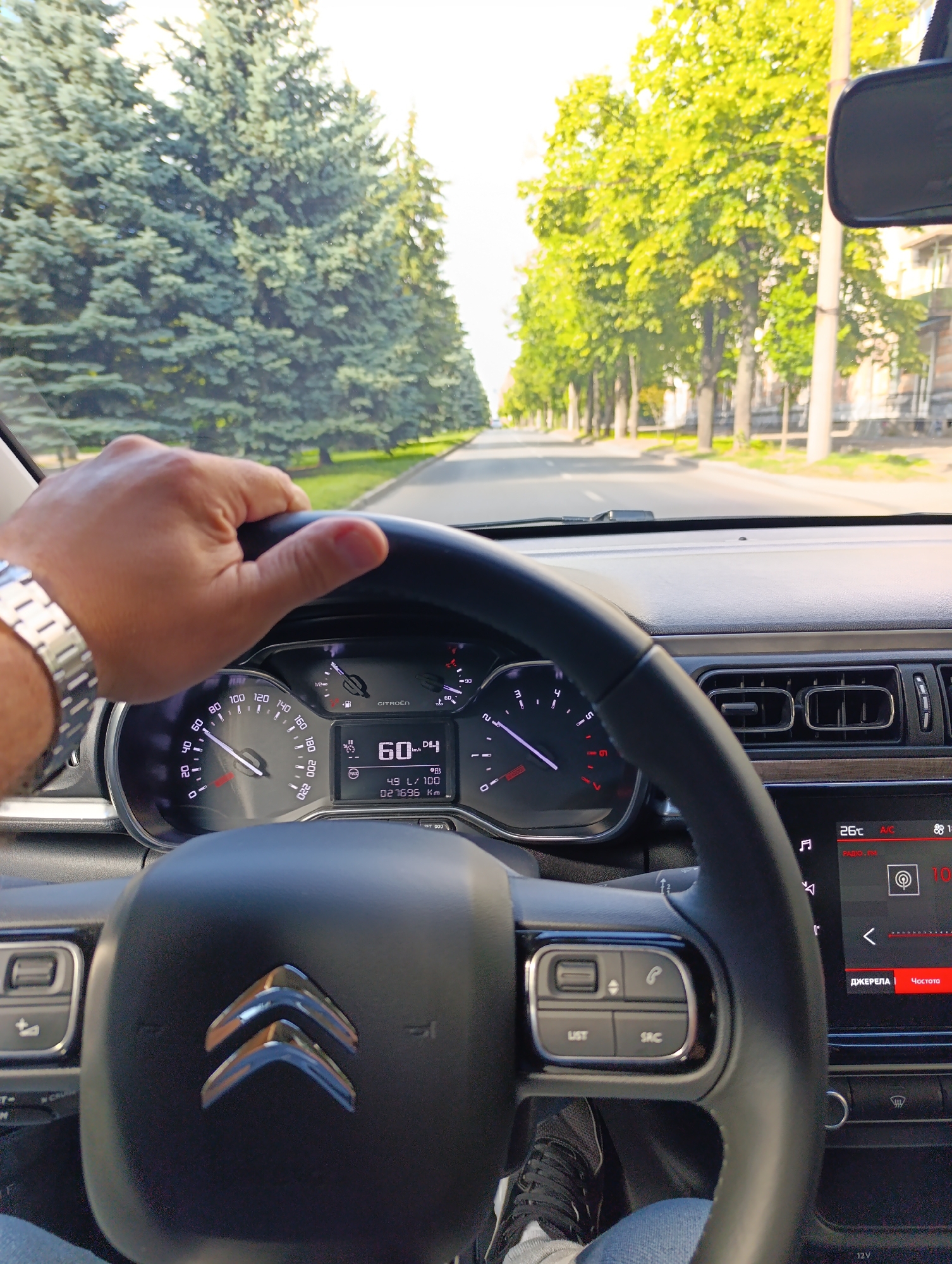 A first-person perspective shot from inside a Citroen vehicle shows the driver's hand on the steering wheel. The dashboard displays the car's speed at 60 km/h and the odometer reading 027696 km. Outside the car, a tree-lined street is visible, with green trees lining both sides of the road. The setting appears to be a residential area in Dnipro, Ukraine.