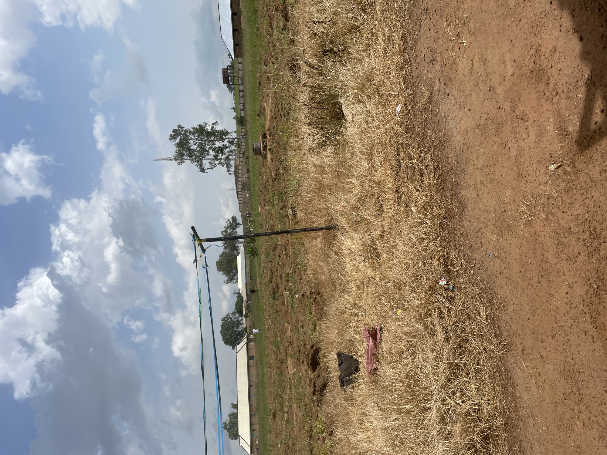 The image depicts an outdoor scene in Durumi, Nigeria. The foreground is dominated by dry, brown grass and a dirt path. In the mid-ground, a black pole with blue and green ribbons or straps attached to it stands vertically. Beyond this, there are buildings with white roofs and darker walls, possibly a school or industrial facility. Some trees are visible in the distance, and a tall, slender structure, possibly a communication tower, is also present. The background consists of a bright, cloudy sky with patches of blue.
There are two people visible in the mid-ground, appearing as small figures. One is wearing white clothing, and another is wearing dark clothing. The overall impression is a daytime scene under a partly cloudy sky. The presence of dry grass and dust suggests a warm, possibly dry season. The scene appears to be a functional area, possibly a school ground or a construction site, given the buildings and the utility pole. There is no visible text in the image.