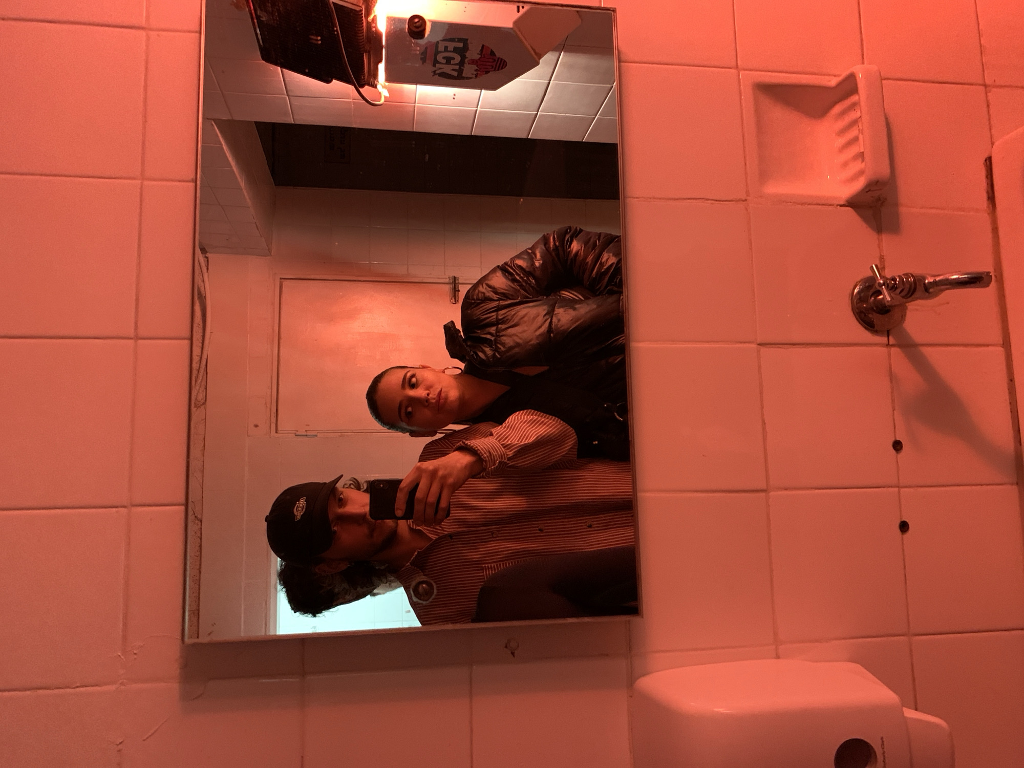 The image displays a reflection of two individuals in a rectangular mirror, located within a restroom facility in Tel-Aviv, Israel. The entire scene is bathed in a pronounced red illumination.
A male subject is reflected on the left, wearing a dark baseball cap featuring a light-colored logo on the front, and a vertical striped shirt. He holds a black smartphone in his right hand, pointed towards the mirror, indicating he is operating the camera. His eyes are visible, directed towards the lens. Dark, curly hair extends from beneath his cap.
Adjacent to him on the right, a female subject is reflected. She is dressed in a shiny, dark puffer jacket and wears large hoop earrings. Her head is angled slightly to her right, with her gaze directed away from the camera. Her facial expression appears neutral.
The background elements visible in the mirror's reflection include a white door that is slightly ajar. To the far right within the reflection, a light source emits red light, and a white wall-mounted dispenser, marked with "FCK" text and a stylized graphic, is present.
The immediate environment outside the mirror's frame consists of white tiled walls. A white soap or sanitizer dispenser with a circular opening is mounted on the wall to the left. Below the mirror, a chrome water faucet with a visible cross-handle extends from the wall, and to its right, a white ceramic soap dish is affixed to the tiled surface. The top edge of a sink basin is partially visible at the bottom of the frame. The overall restroom fixtures and tiling appear functional.