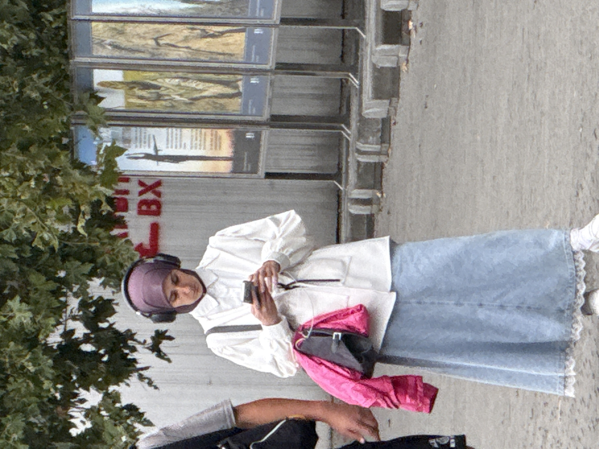 A person wearing a purple hijab and headphones stands in the foreground of an urban scene in Inkerman, Ukraine. They are dressed in a white collared shirt, a light blue denim skirt with white lace trim, and white sneakers. A black handbag with a pink jacket draped over it hangs from their shoulder. The individual is looking down at a smartphone held in their hands. To the left, another person, partially visible, wears a dark grey t-shirt and a black backpack. Behind them, a concrete wall with red lettering reads "BX". A row of tall display panels with images and text stands in the background, each mounted on a concrete base. Green foliage from a tree overhangs the scene from above. The ground is paved with asphalt.