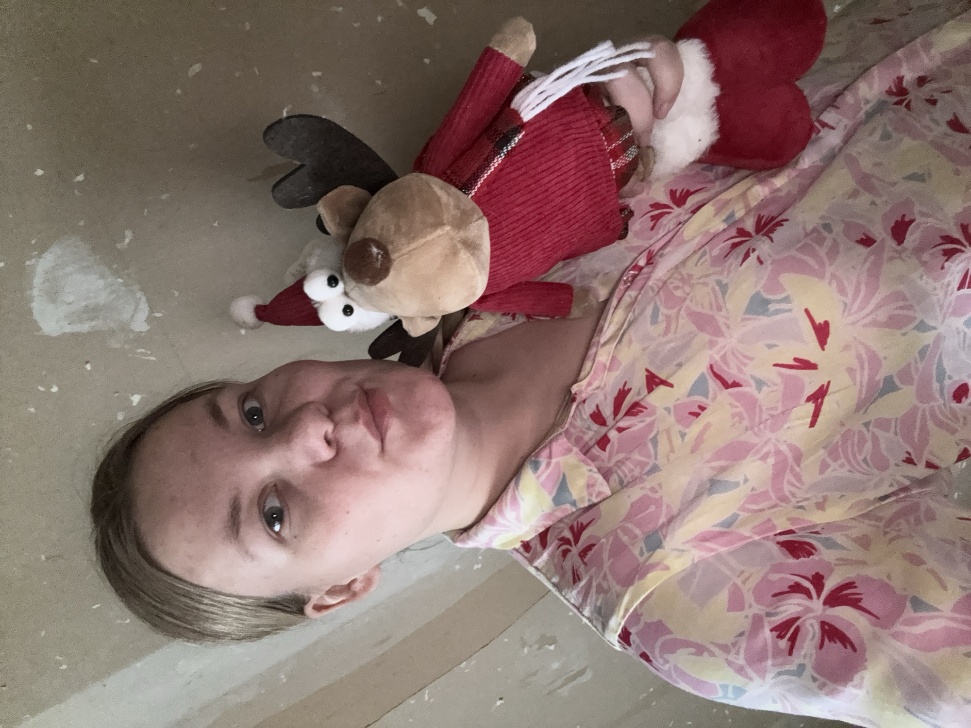 An overhead portrait-style image, originally rotated 90 degrees counter-clockwise, features a young person lying down, holding a festive stuffed animal. The person, with light-colored hair and eyes, is looking directly at the camera with a neutral to slightly pouting expression.
The primary subject is a young individual, likely female, whose head and upper torso are visible. She is wearing a patterned garment, possibly pajamas or a casual top, adorned with pink, red, yellow, and light blue floral or leaf designs.
Resting beside or on her is a plush reindeer toy. The reindeer has a brown body, dark antlers, prominent white eyes, and is wearing a red Santa hat with a white brim and pompom. It also sports a red knitted sweater and a plaid scarf with red, black, and white patterns. The person's hand is visible, gently holding a part of the reindeer or its red stocking-like arm.
The background appears to be a light, possibly off-white or beige surface, such as a floor or wall, with scattered white specks that could be dust, paint flakes, or debris, suggesting an unfinished or simple indoor setting. There are no clear indications of the time of day, weather, or specific room type. No visible text is present in the image. The location context provided is Chaikovsky, Russia.