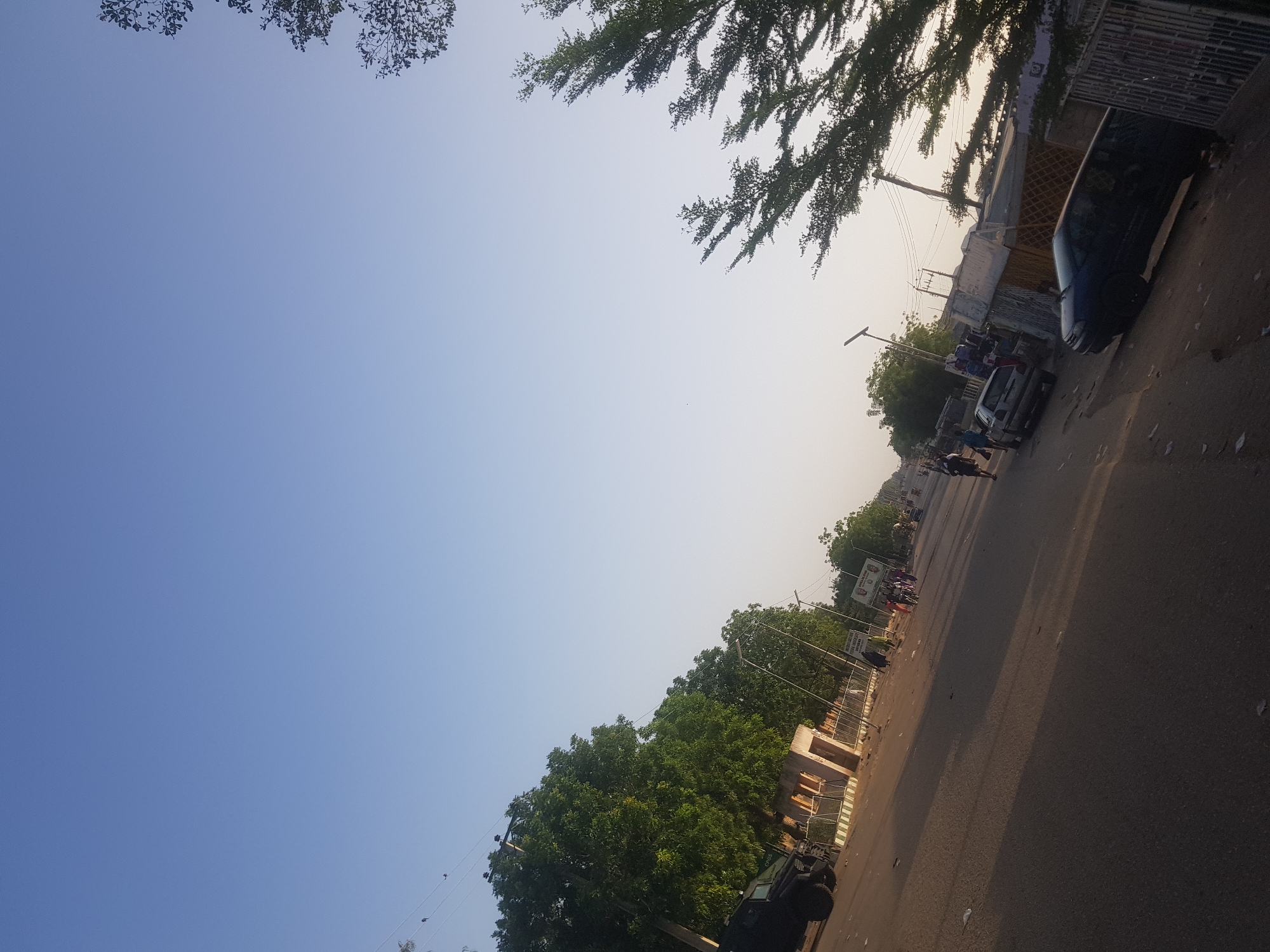 The image shows a street scene in Dabawa, Nigeria. The sky is clear and blue with sparse tree branches visible in the upper corners. A paved road stretches into the distance, with cars and pedestrians visible along it. Buildings and trees line both sides of the road. A dark colored vehicle is parked on the right side. Light poles stand along the road. The light suggests it's either morning or late afternoon.