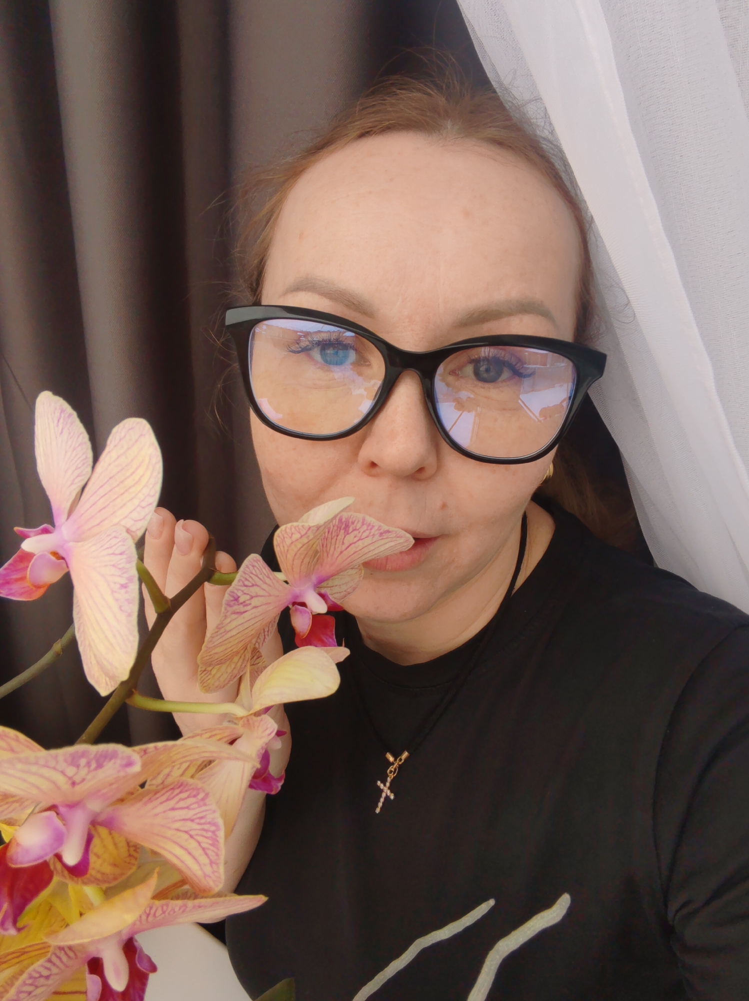 A woman with reddish-blonde hair, wearing large black-framed glasses, a black t-shirt, and a gold cross necklace, poses with a stem of striped orchids. She holds the flower near her face, almost as if kissing it. The background includes a dark brown curtain and a partially visible white curtain. The image was taken in the city of Chaikovsky, Russia.