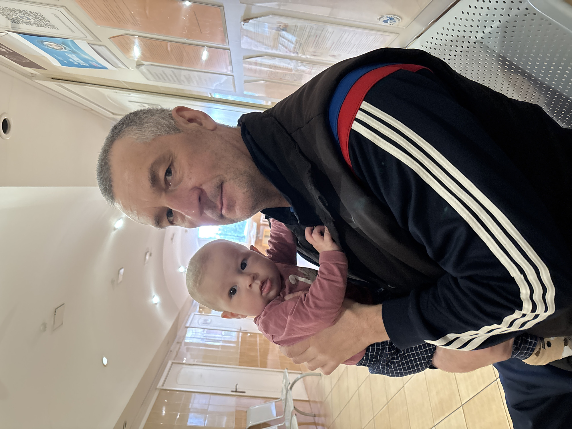 A man is holding a baby in an indoor location that appears to be a waiting area of a medical facility, potentially in Sochi, Russia. The man has gray hair and is wearing a dark jacket with white stripes on the sleeves, along with a blue and red stripe around the neck. The baby is wearing a light pink long-sleeved shirt and patterned pants. The background shows light-colored walls with recessed lights, and there are signs and notices on the wall near the ceiling. In the distance, there are rows of chairs, which are typical of waiting rooms.