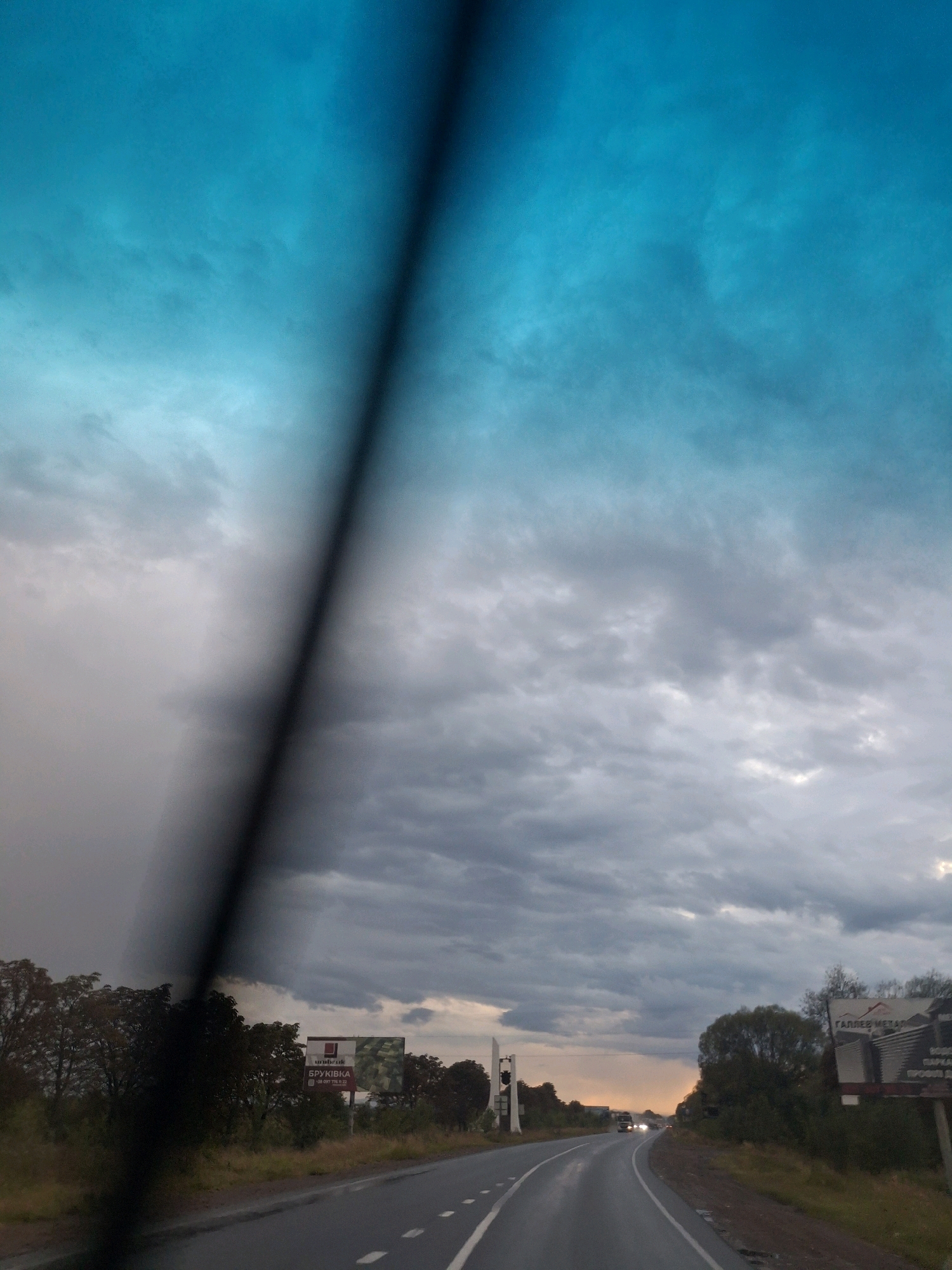 The image depicts a rural roadside scene in the city of Piilo, Ukraine, under a dramatic, cloudy sky. The perspective is from inside a vehicle, with a blurred black vertical streak across the left portion of the frame, possibly a windshield wiper or a reflection. The sky is a striking gradient of deep blue and turquoise at the top, transitioning to heavy, grey storm clouds lower down.
The road curves to the right, appearing wet, indicating recent rain. The road surface is asphalt with white lane markings. On the right side of the road, there are trees and dense vegetation. A large billboard with text in Cyrillic script and an image of what appears to be a building material is visible. The text on the billboard appears to be "UNIBROK BRUSSKIVKA" and a phone number. Further down the road, in the distance, other vehicles can be seen, including headlights suggesting traffic. On the left side of the road, there are more trees and a few distant structures. The overall atmosphere suggests a late afternoon or early evening with impending or recent rainfall.