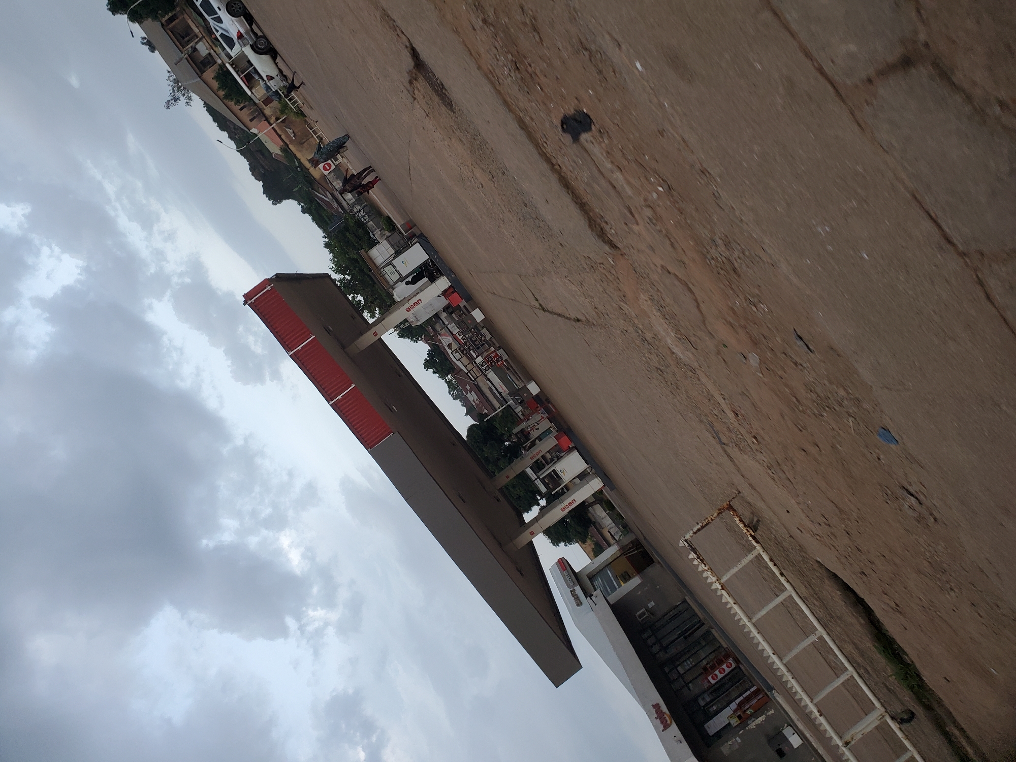 The image depicts a gas station in Bukuru, Nigeria, under an overcast sky. The main subject is a modern gas station with a prominent red and gray canopy. Several fuel pumps are visible under the canopy, some bearing the "Shell" logo. Adjacent to the pumps is a building with large glass windows, likely a convenience store, displaying branding that includes "Harbour" and "Go-Kart." In the background, there are more buildings, some appearing weathered, and lush green trees. A white SUV is parked near the buildings, and a few individuals, including what appears to be a family group, are walking on the asphalt. The ground is a mix of asphalt and dirt, with visible cracks and patches. A white metal barrier with a saw-tooth edge is positioned in the foreground, angled across the frame. The overall impression is that of a typical, functional gas station in a semi-urban or town setting.